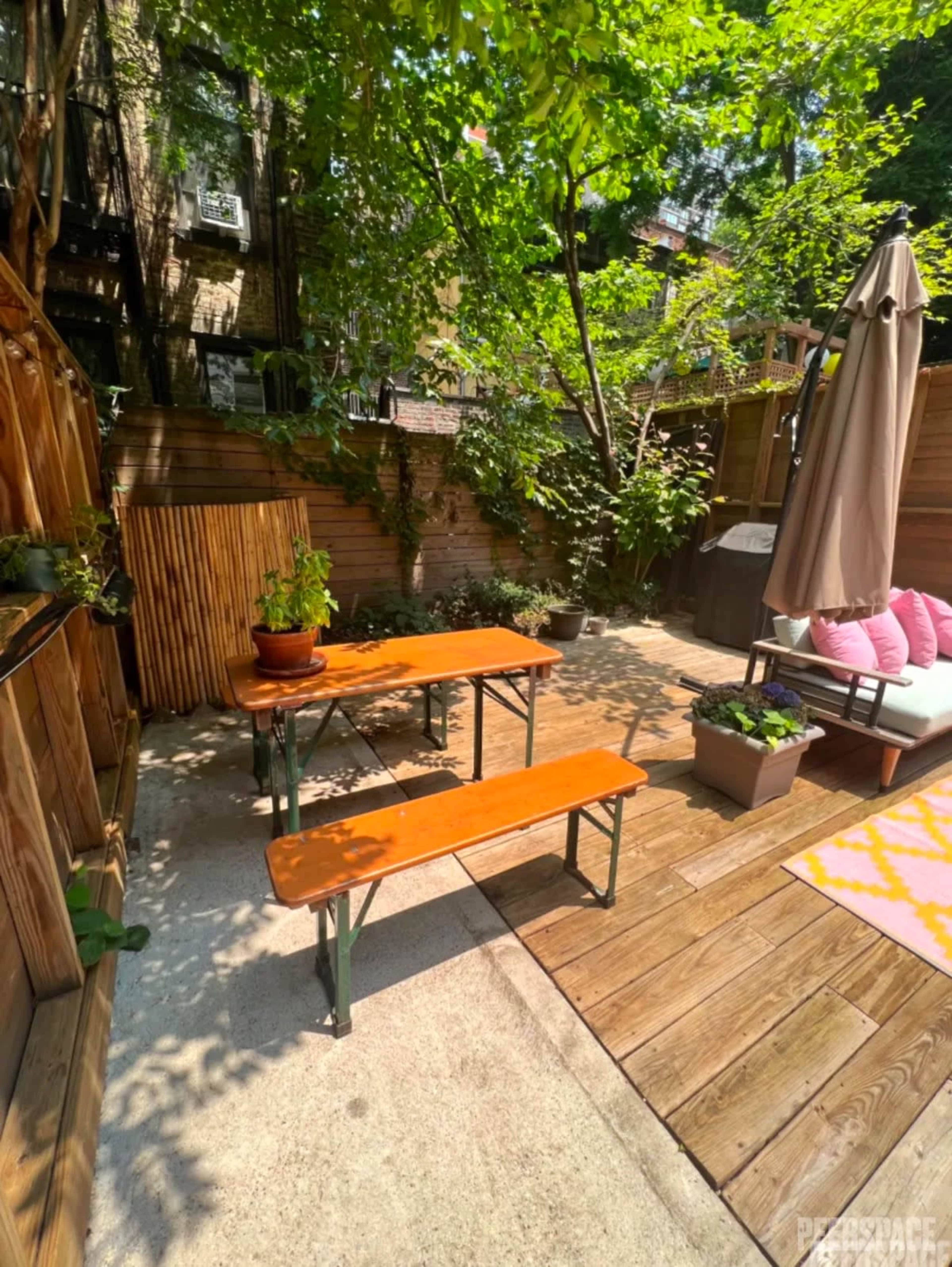 A small outdoor patio features a wooden table and benches surrounded by greenery and potted plants.