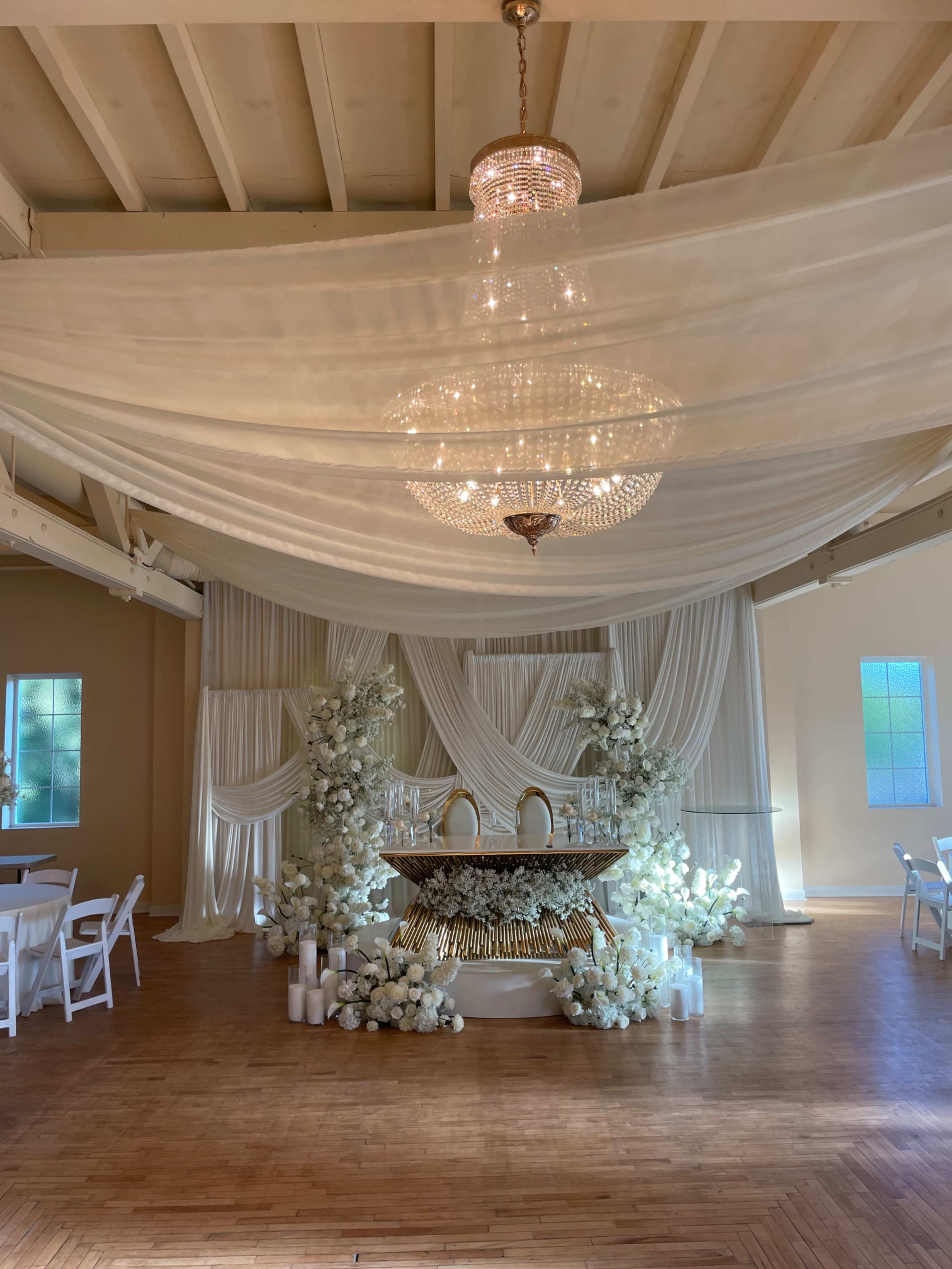 The image shows an elegantly decorated venue featuring a chandelier, draped white fabric, and floral arrangements surrounding a central table.