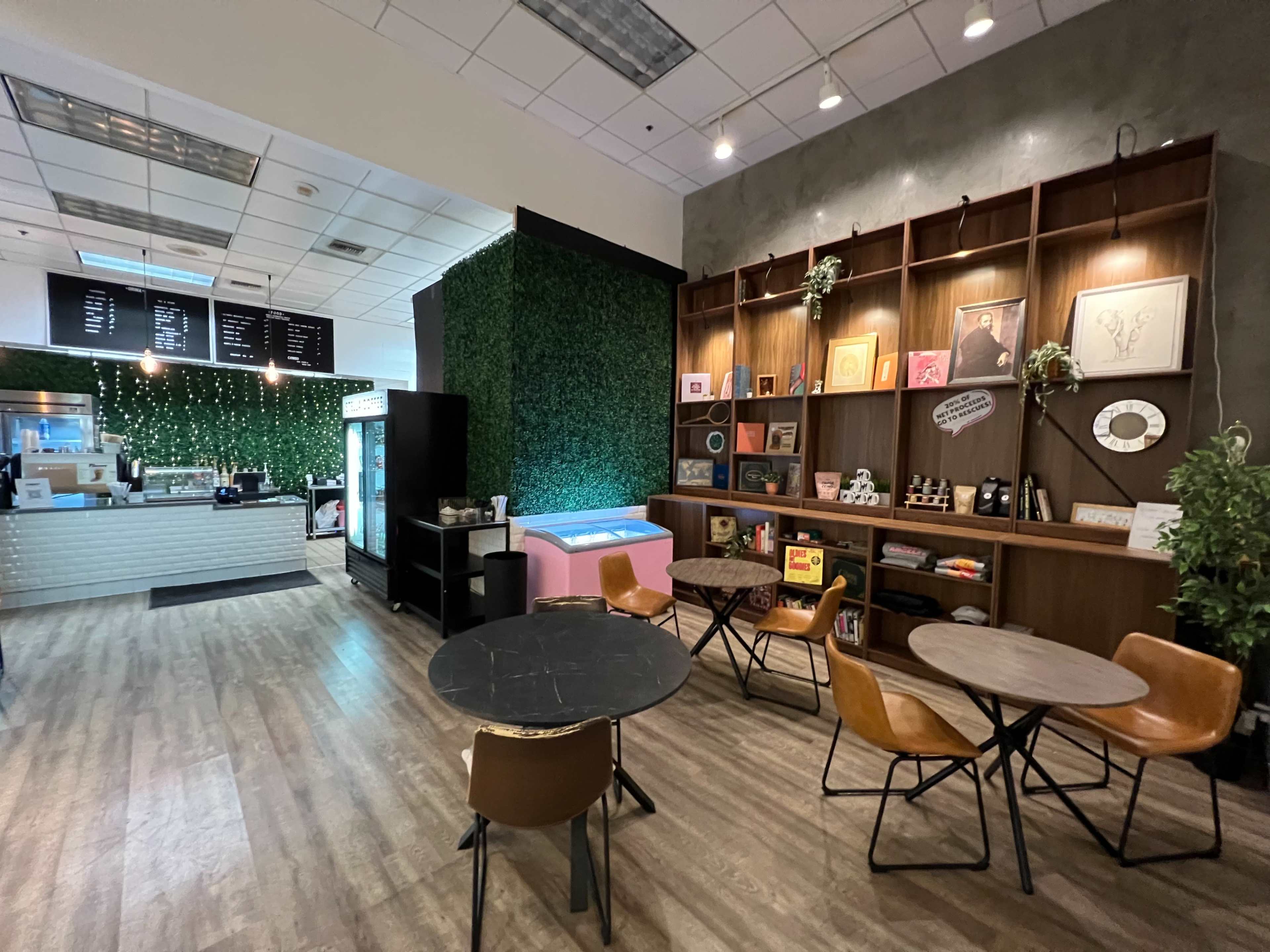 The image shows a modern cafe interior with a coffee counter, seating area featuring wooden tables and chairs, and decorative wall shelves displaying various framed items and plants.