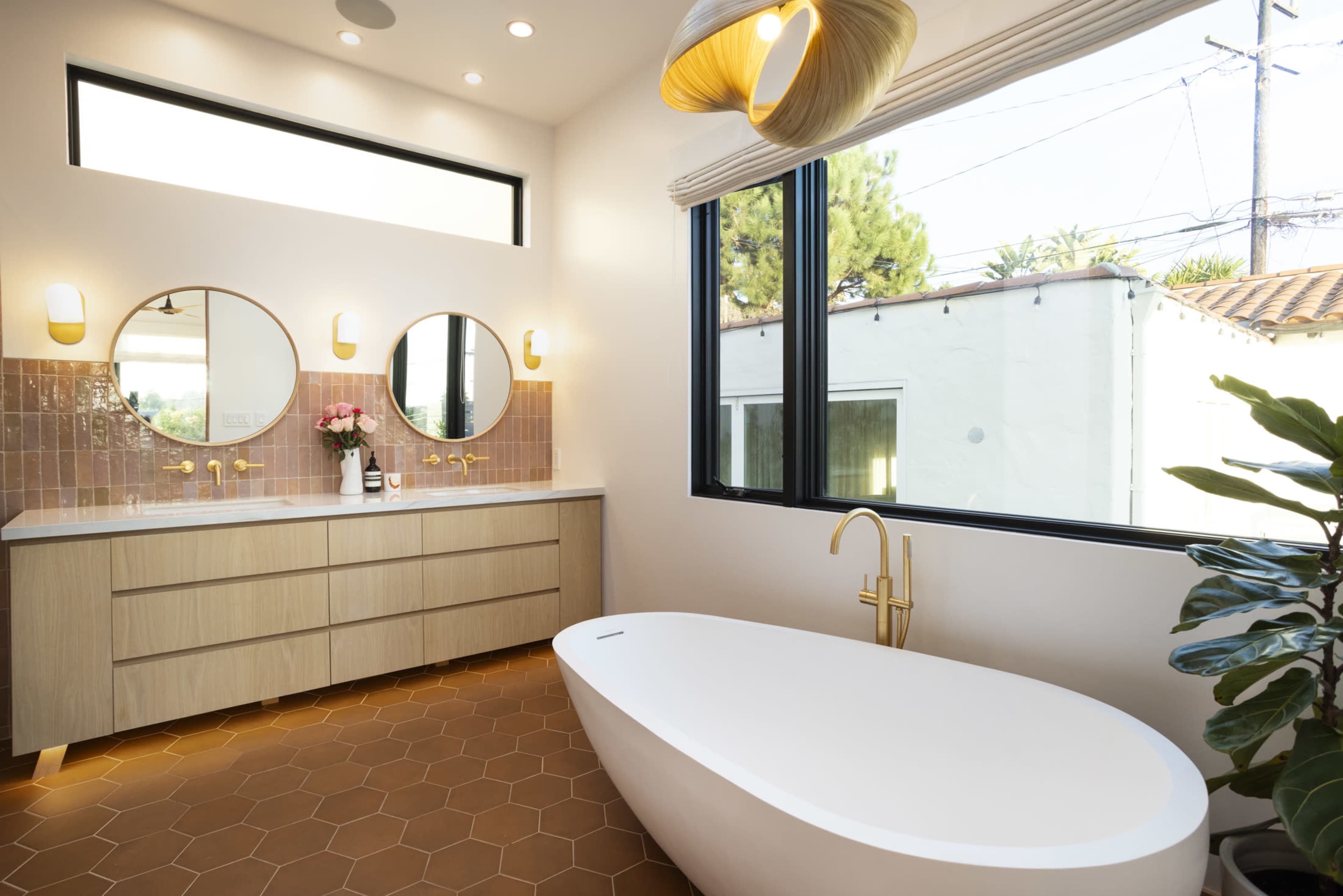 A modern bathroom features a freestanding bathtub, dual round mirrors, and a wooden vanity with gold fixtures, illuminated by natural light from large windows.