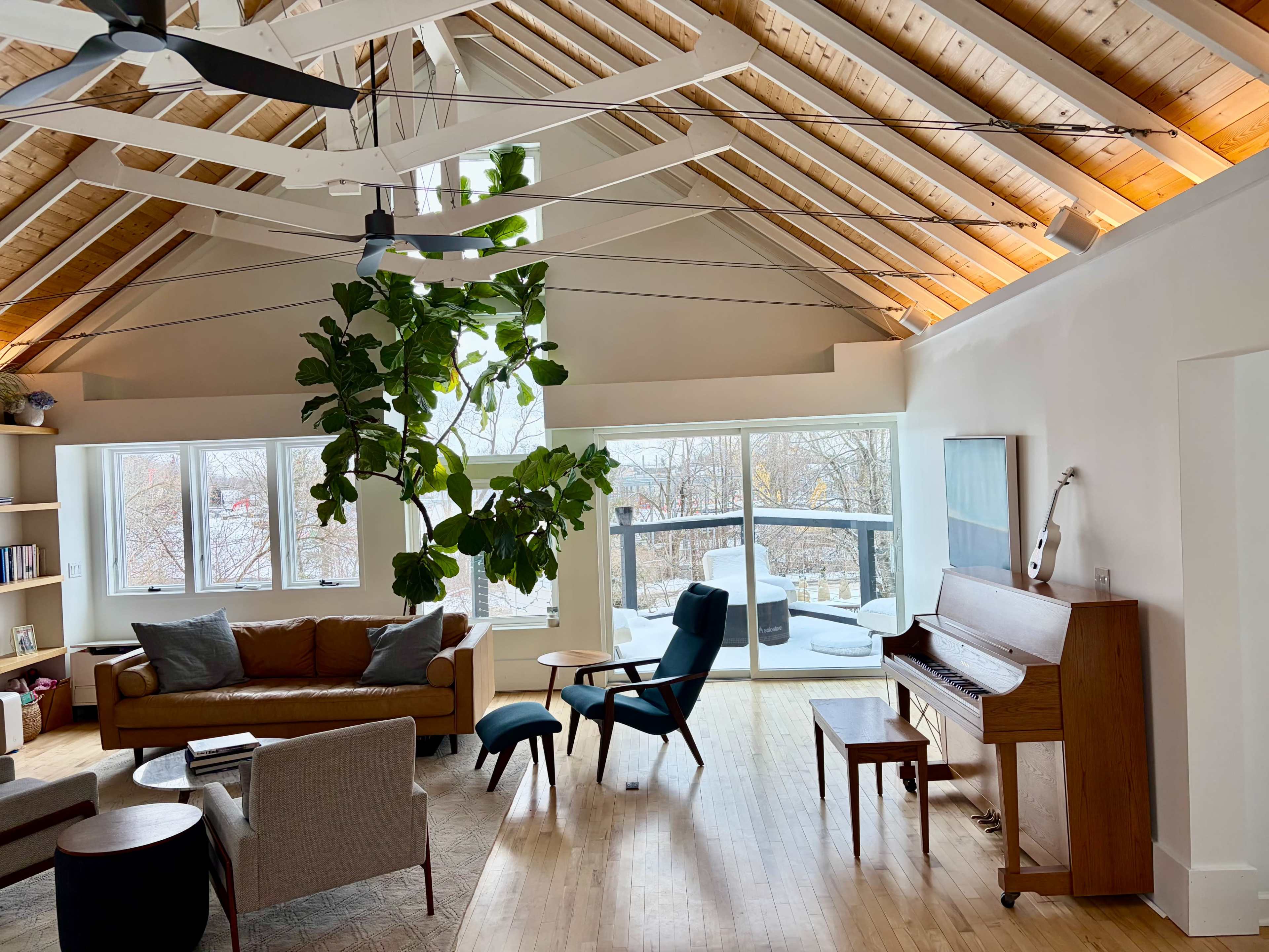 The room features a spacious living area with a wooden ceiling, a large window, a piano, and a tall indoor plant.