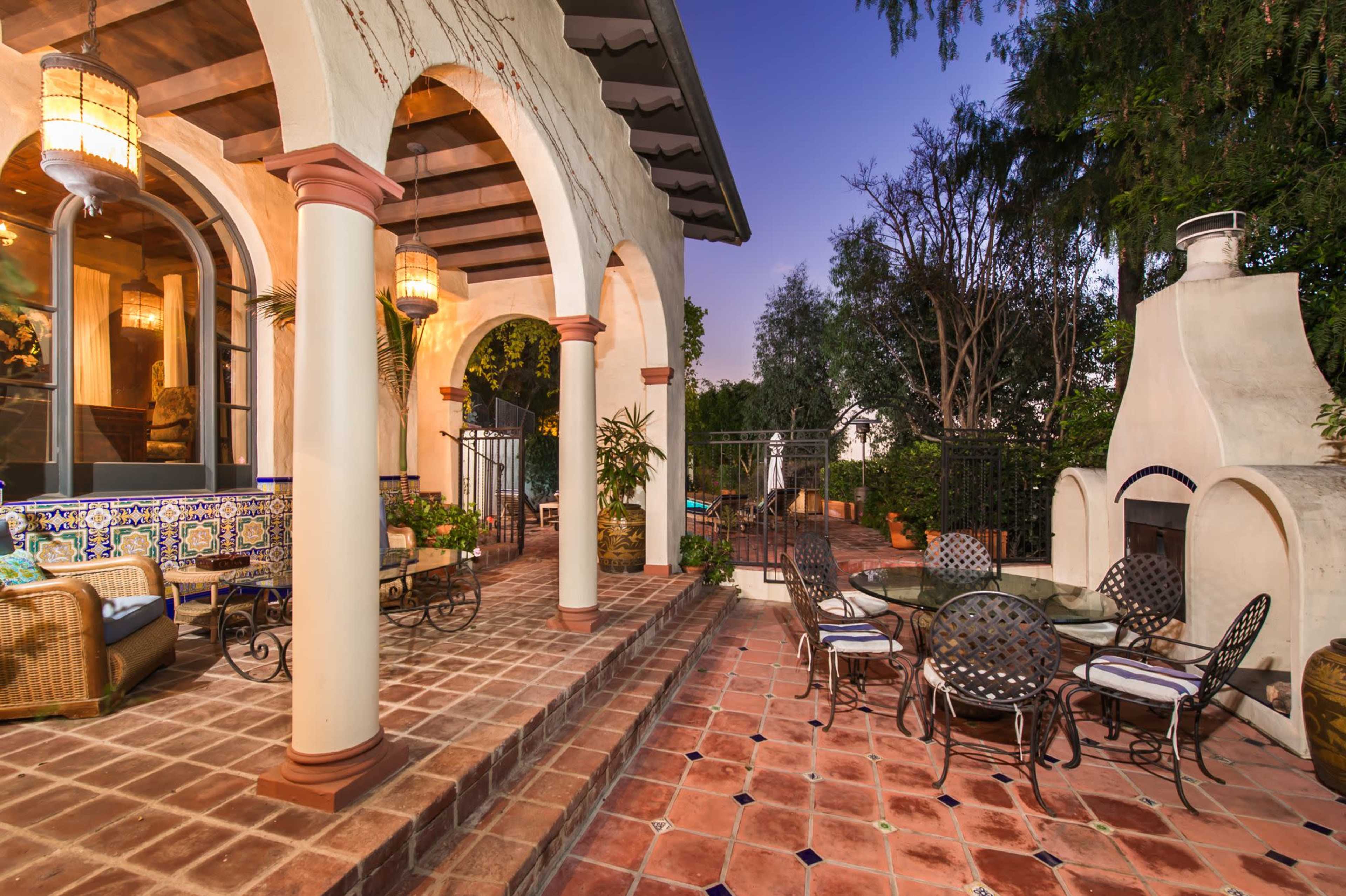A patio area featuring tiled flooring, arches supported by columns, and a fireplace beside a swimming pool, surrounded by greenery.