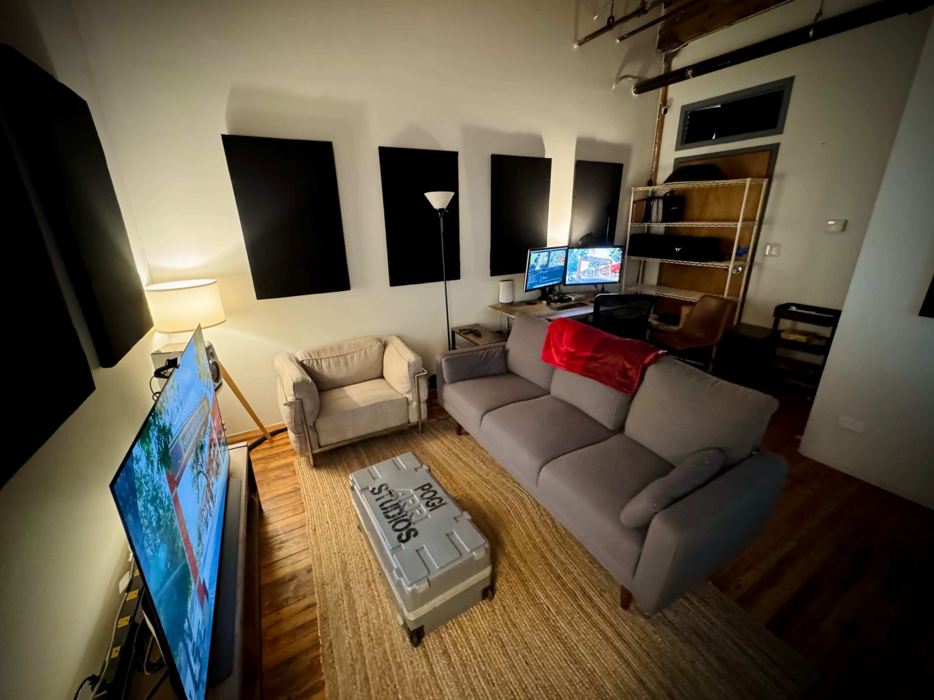The image shows a cozy room with a gray sofa, a light-colored armchair, a large television, and two computer monitors on a desk against the wall.