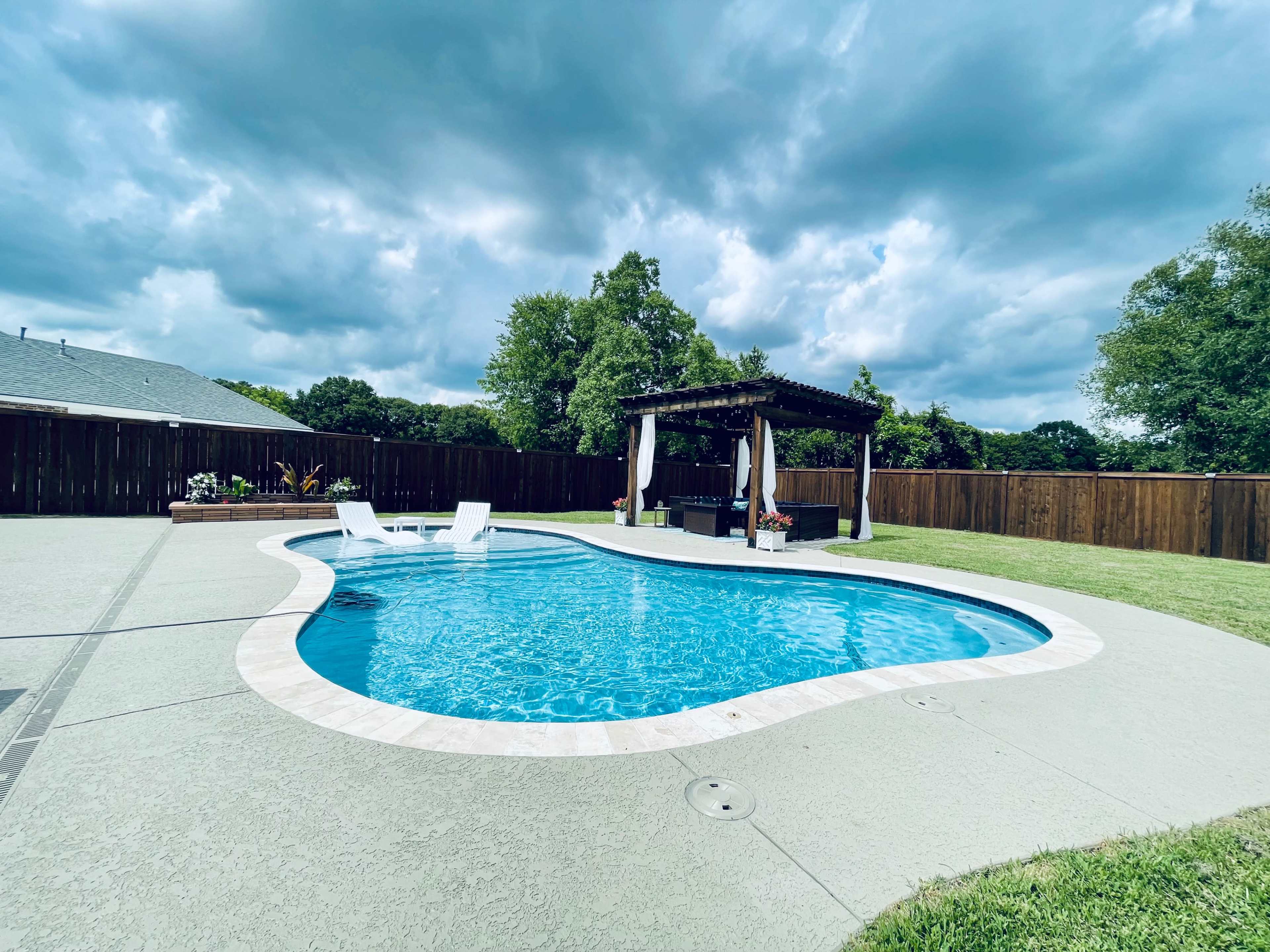A swimming pool with a curved shape is surrounded by a patio and a gazebo, set against a cloudy sky in a backyard.