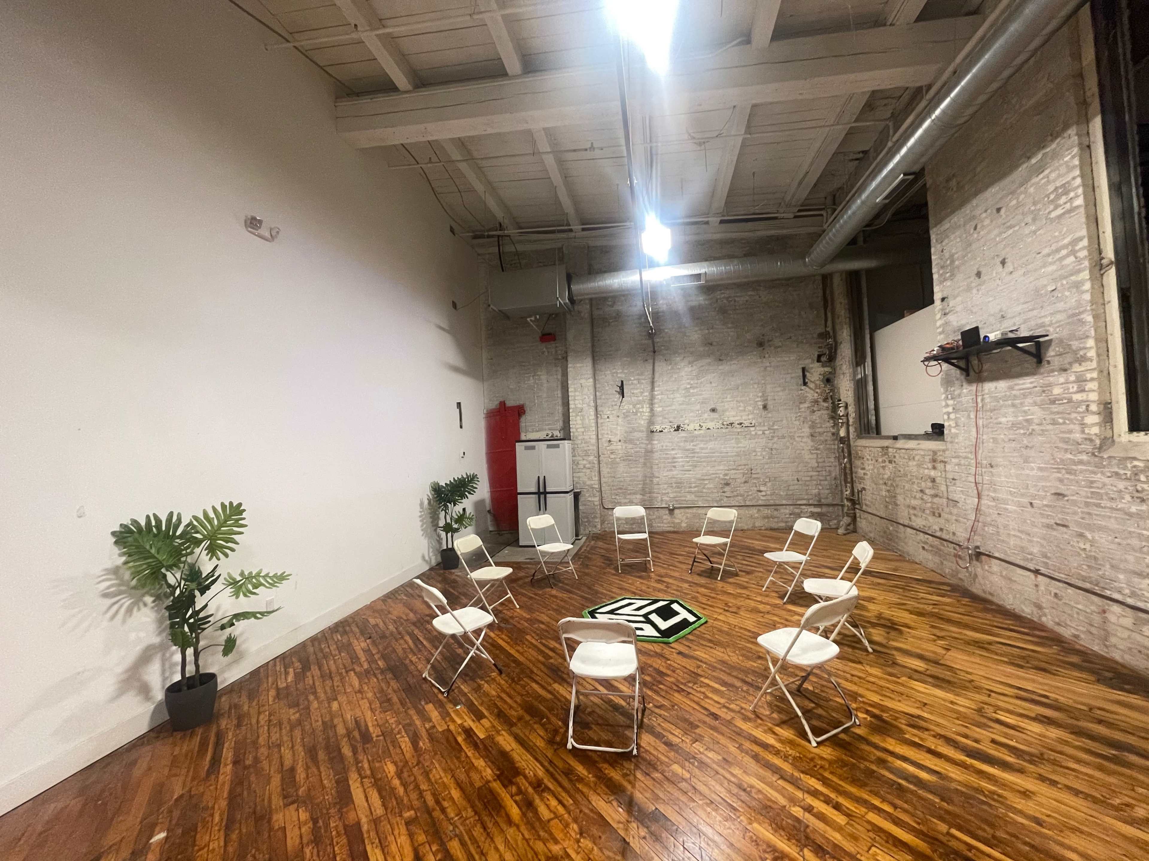 The image shows a spacious room with wooden flooring, featuring a circle of white folding chairs arranged around a black and white patterned mat, with a small plant in a pot on one side.