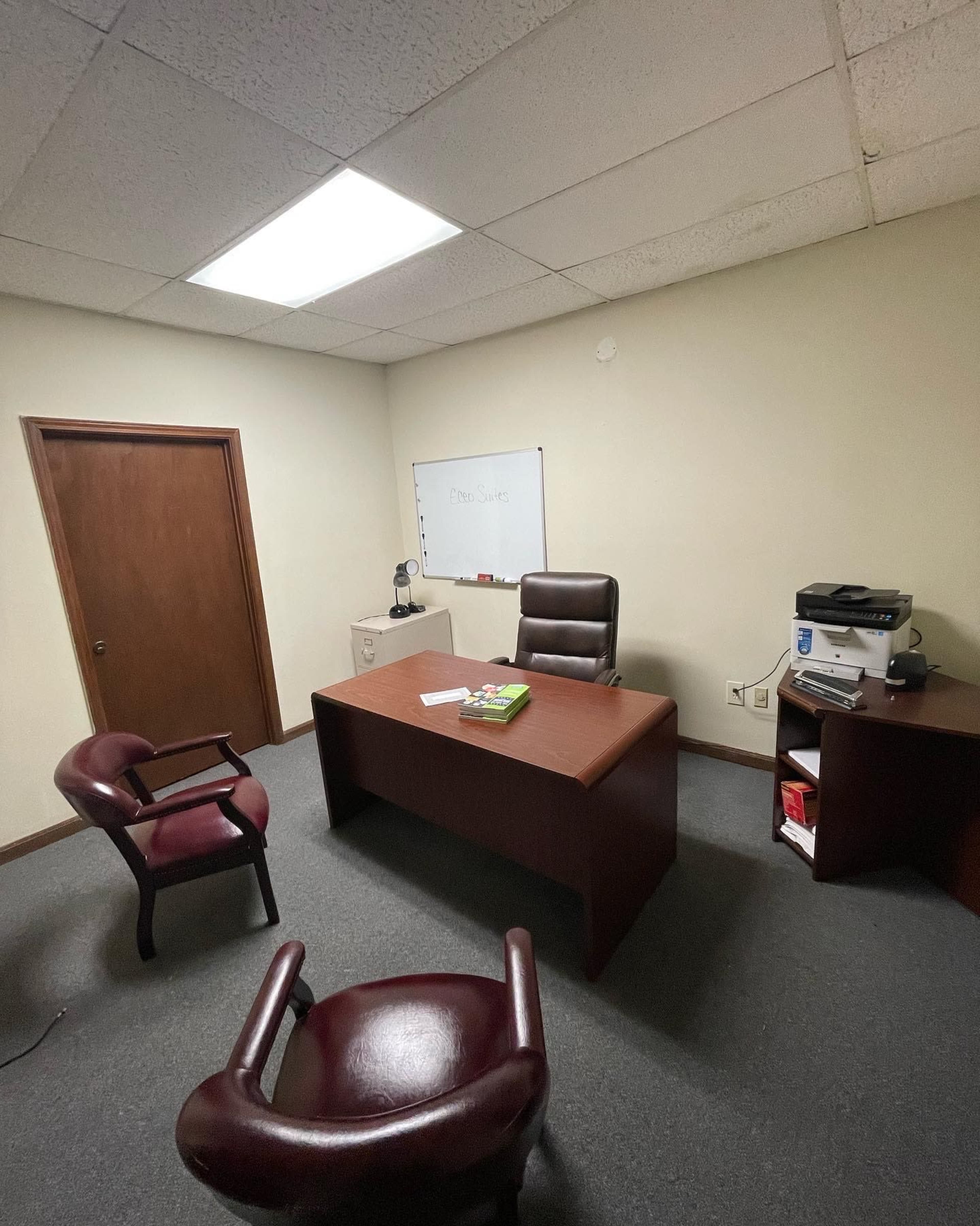 A sparsely furnished office with a wooden desk, an office chair, two chairs in front, a whiteboard on the wall, and a printer on a side table.