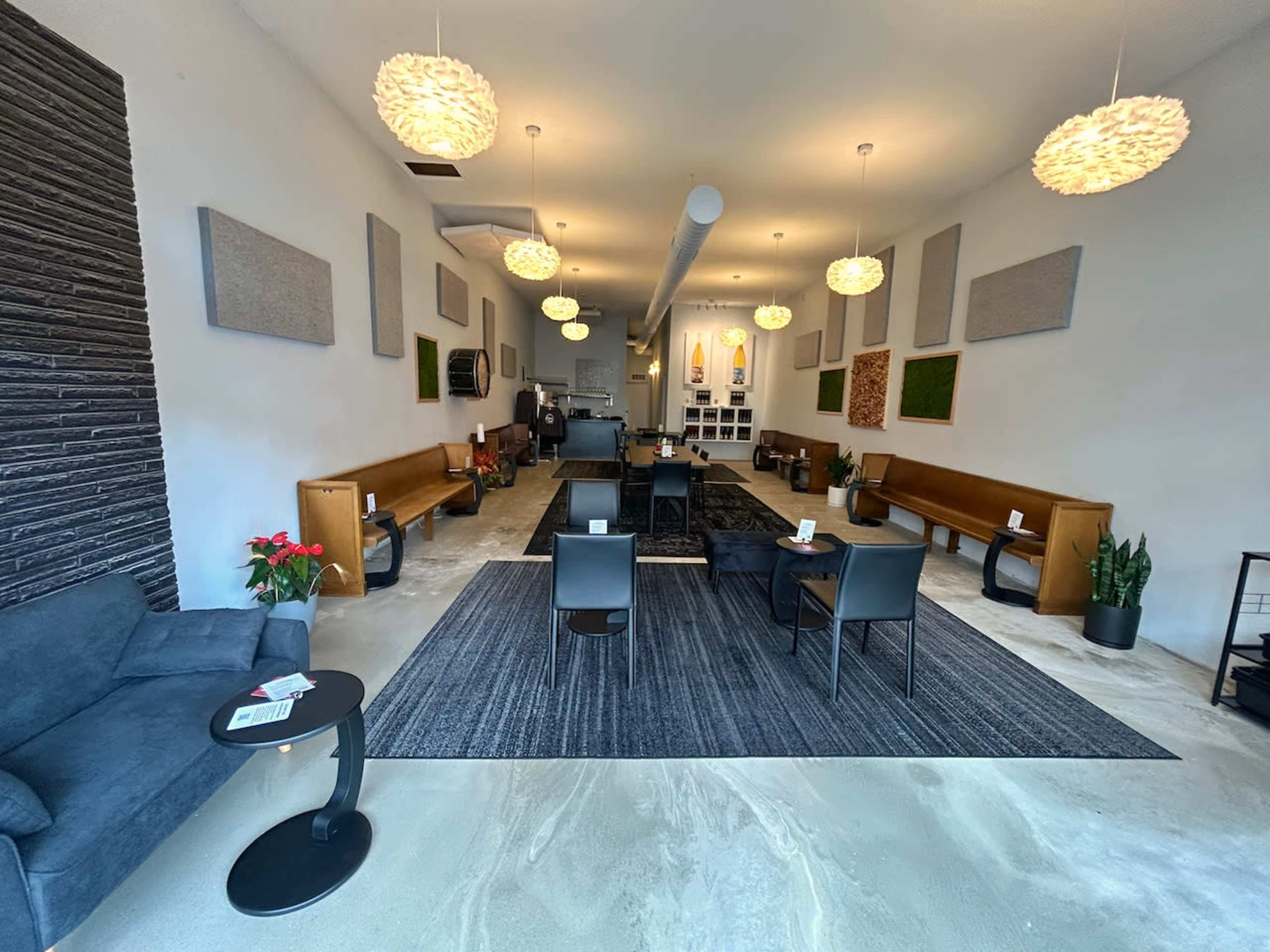 A spacious interior of a minimally decorated lounge with wooden benches, black chairs, and plants, along with pendant lighting and sound-absorbing panels on the walls.