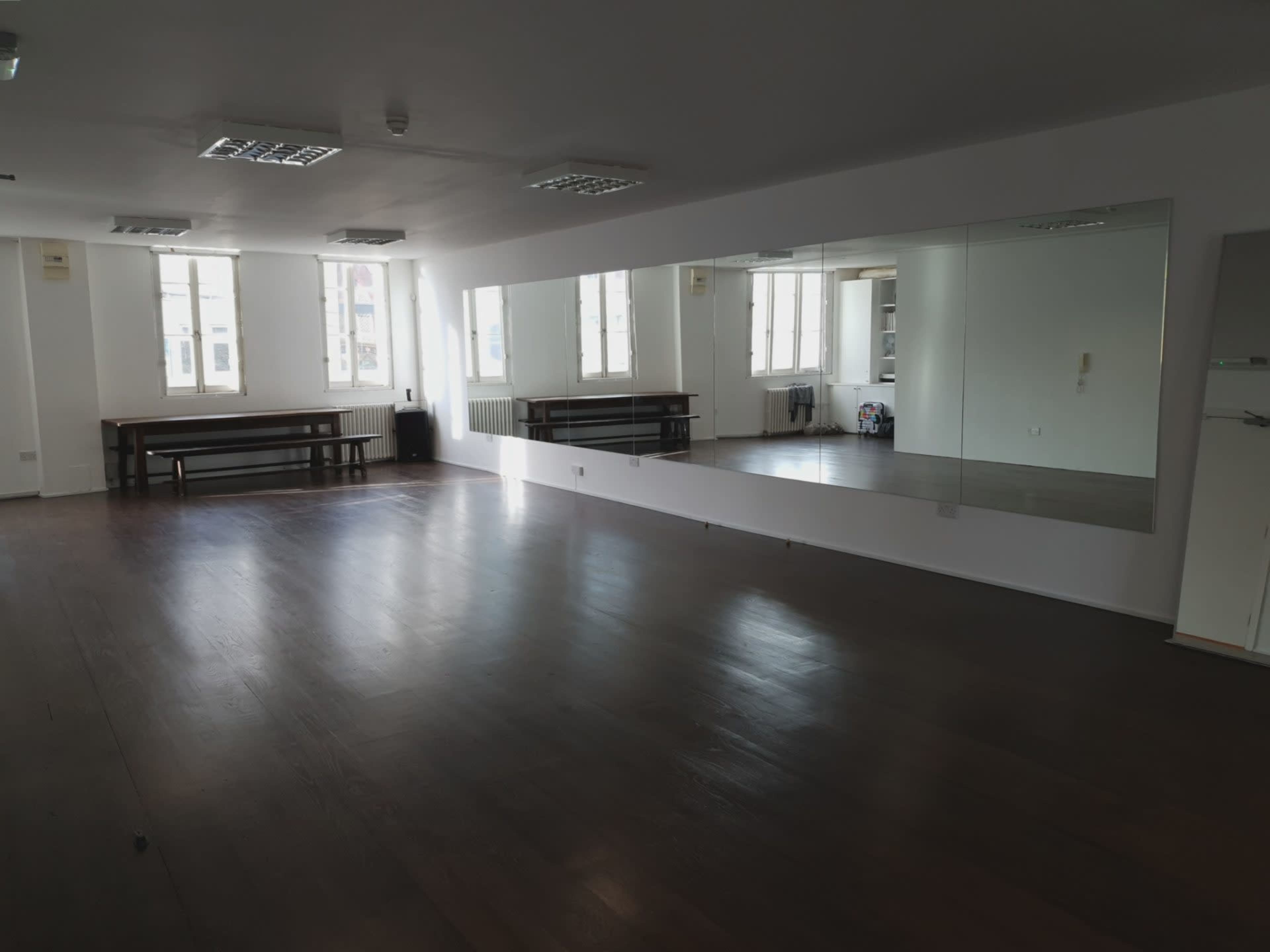 An empty room with wooden floors, large windows, and a long mirror along one wall.