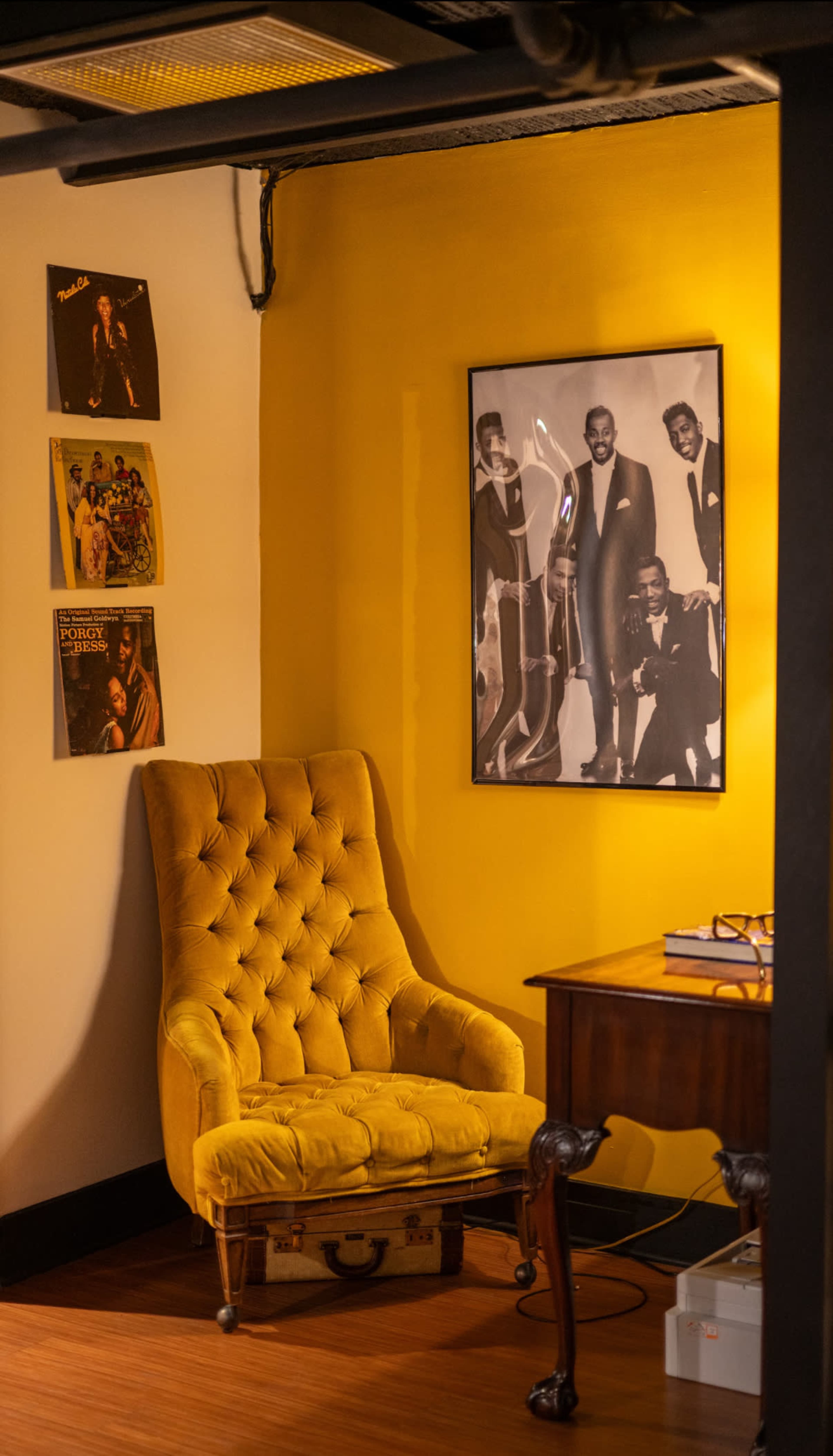 The image shows a yellow-walled corner of a room featuring a tufted yellow chair, a wooden desk, and framed black-and-white photographs on the wall.