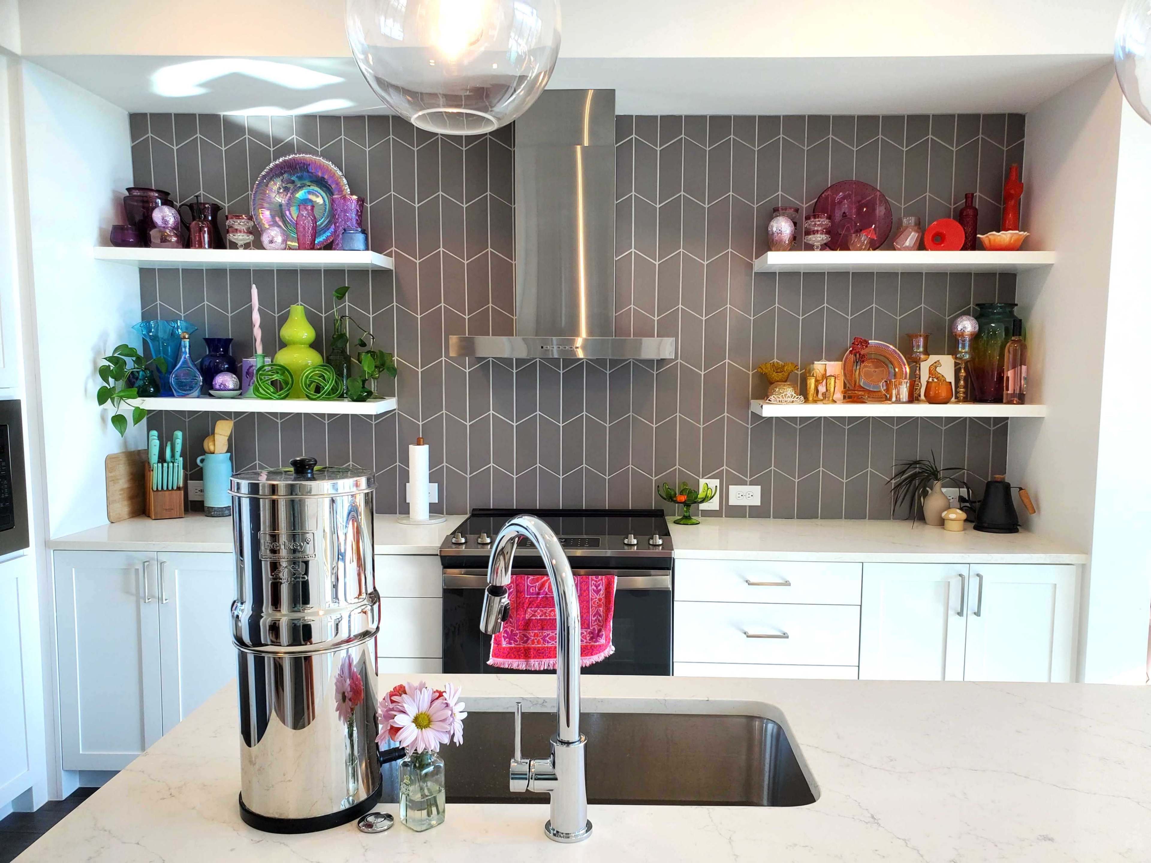 The modern kitchen features a marble countertop, a stainless steel sink, a water filter, and colorful glassware neatly arranged on open shelves against a patterned gray backsplash.
