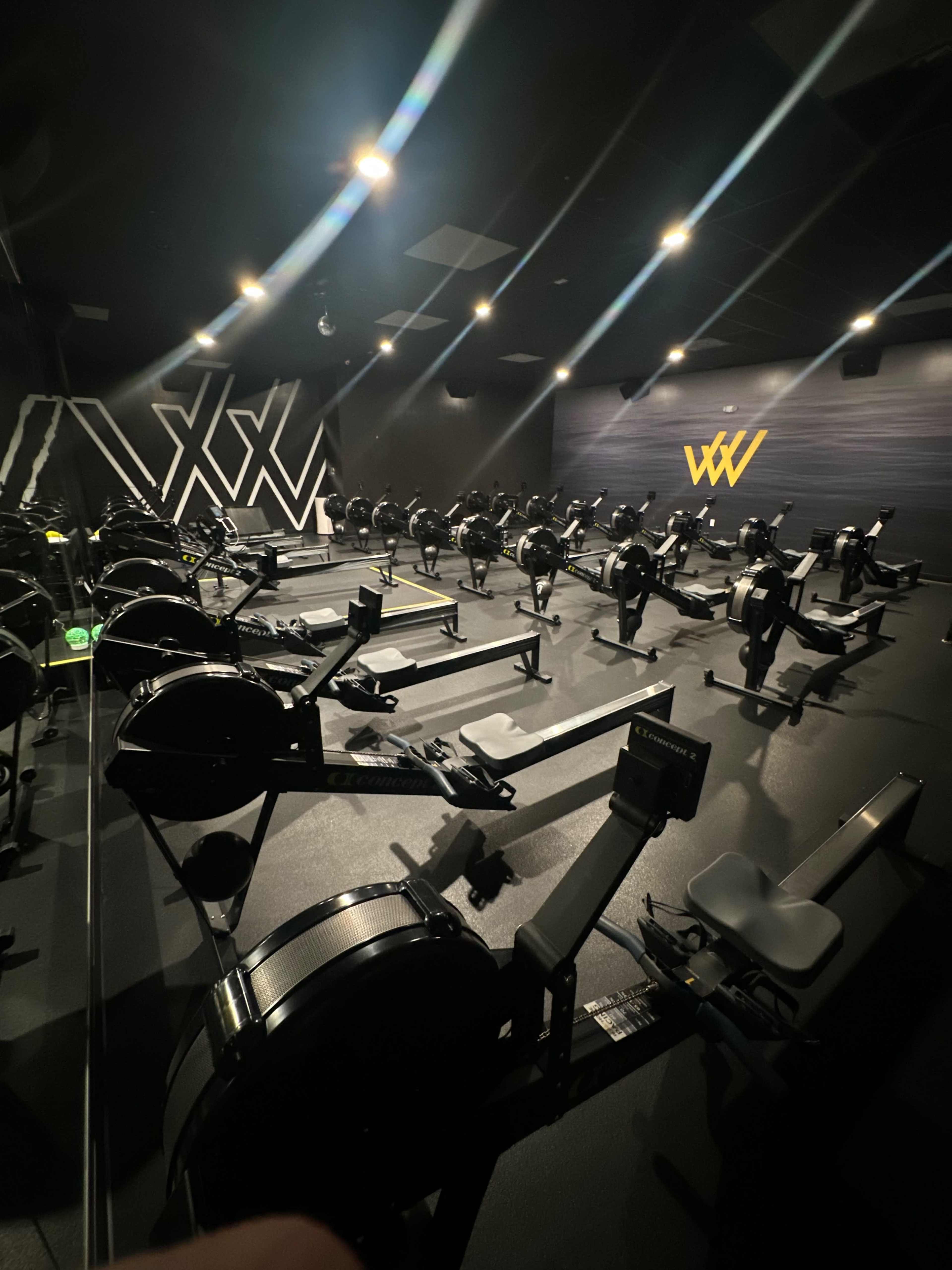 The image shows an indoor rowing gym equipped with multiple rowing machines and a modern, minimalist design.