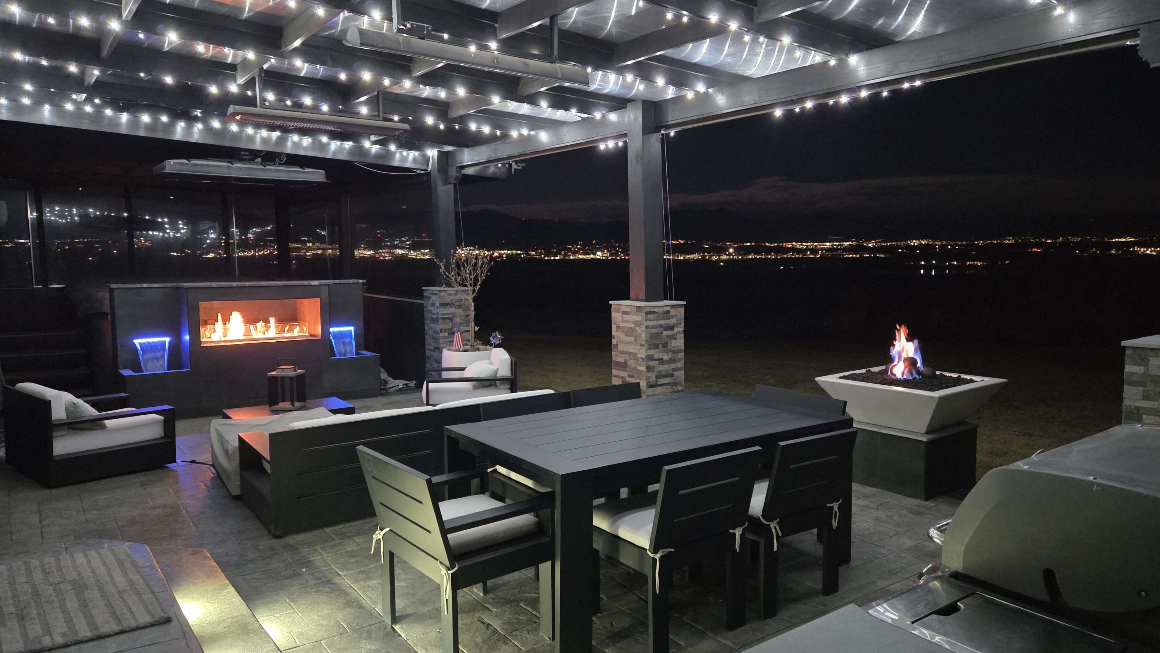 The image shows a modern outdoor patio with a dining table, seating area, and a fire feature, illuminated by string lights against a night sky.