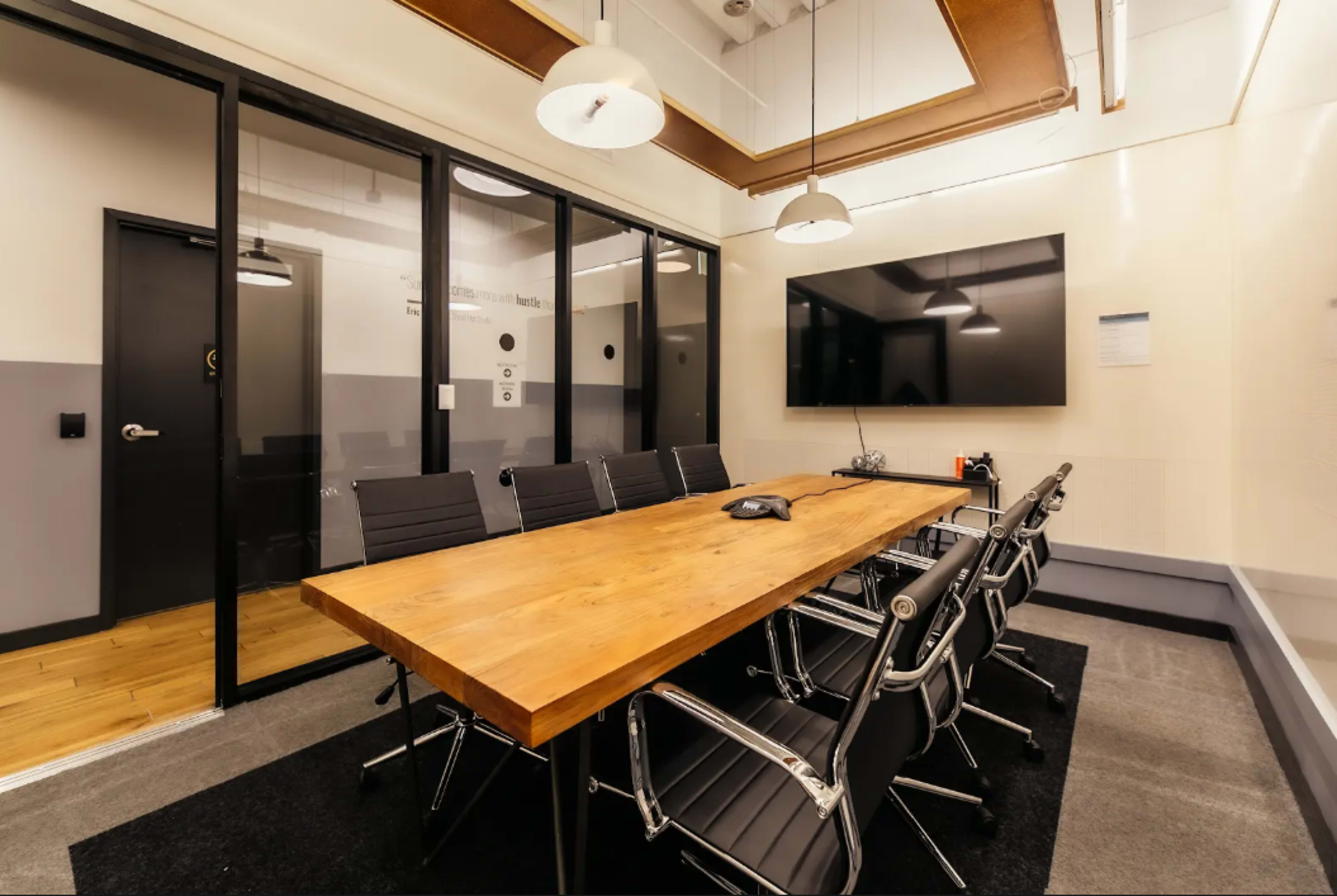 A modern conference room features a long wooden table surrounded by sleek black chairs and equipped with a large display screen.