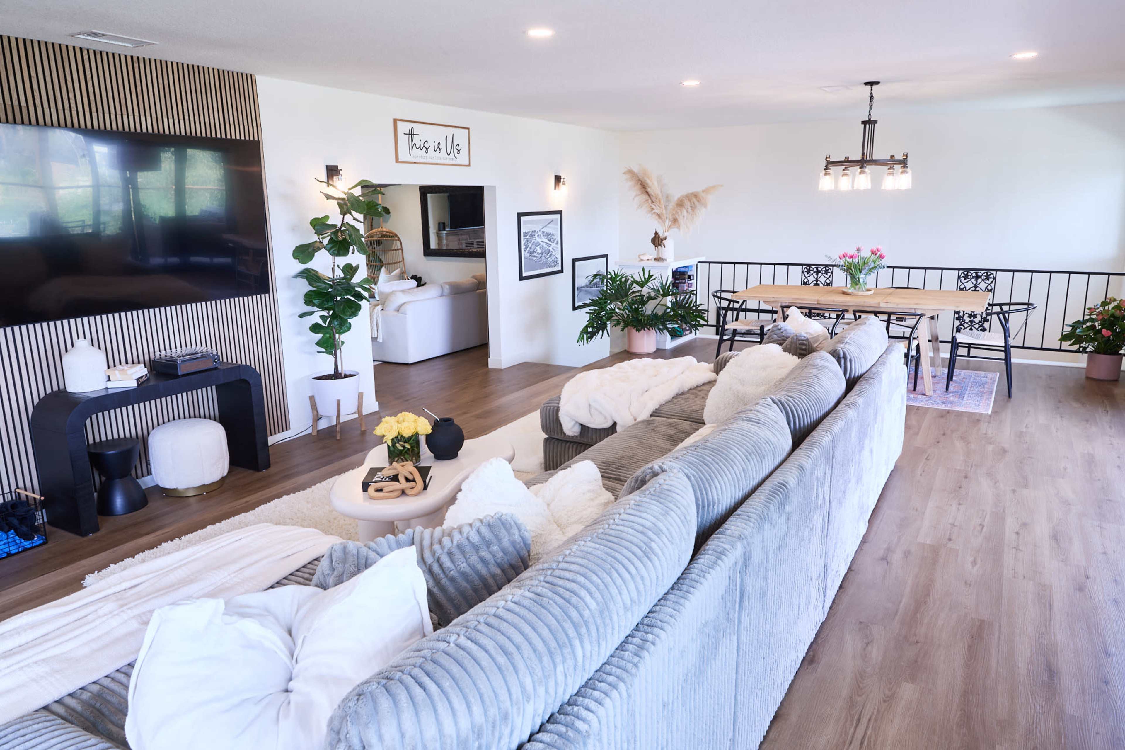 A modern living space with a large, upholstered sectional sofa facing a TV, surrounded by plants and a dining area with a wooden table and black chairs.
