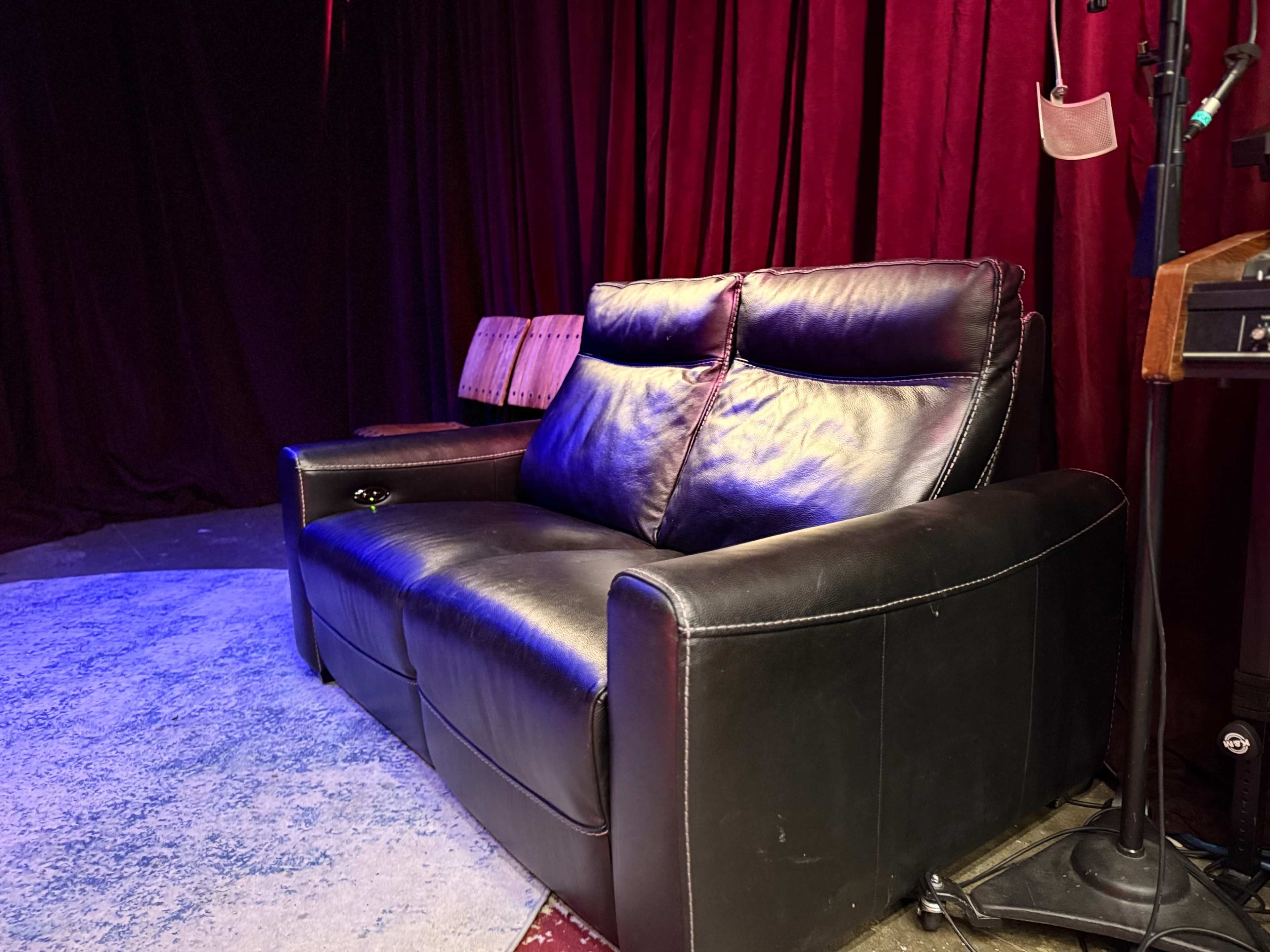A black leather couch is positioned in front of a red curtain, with a small table and chairs visible in the background.
