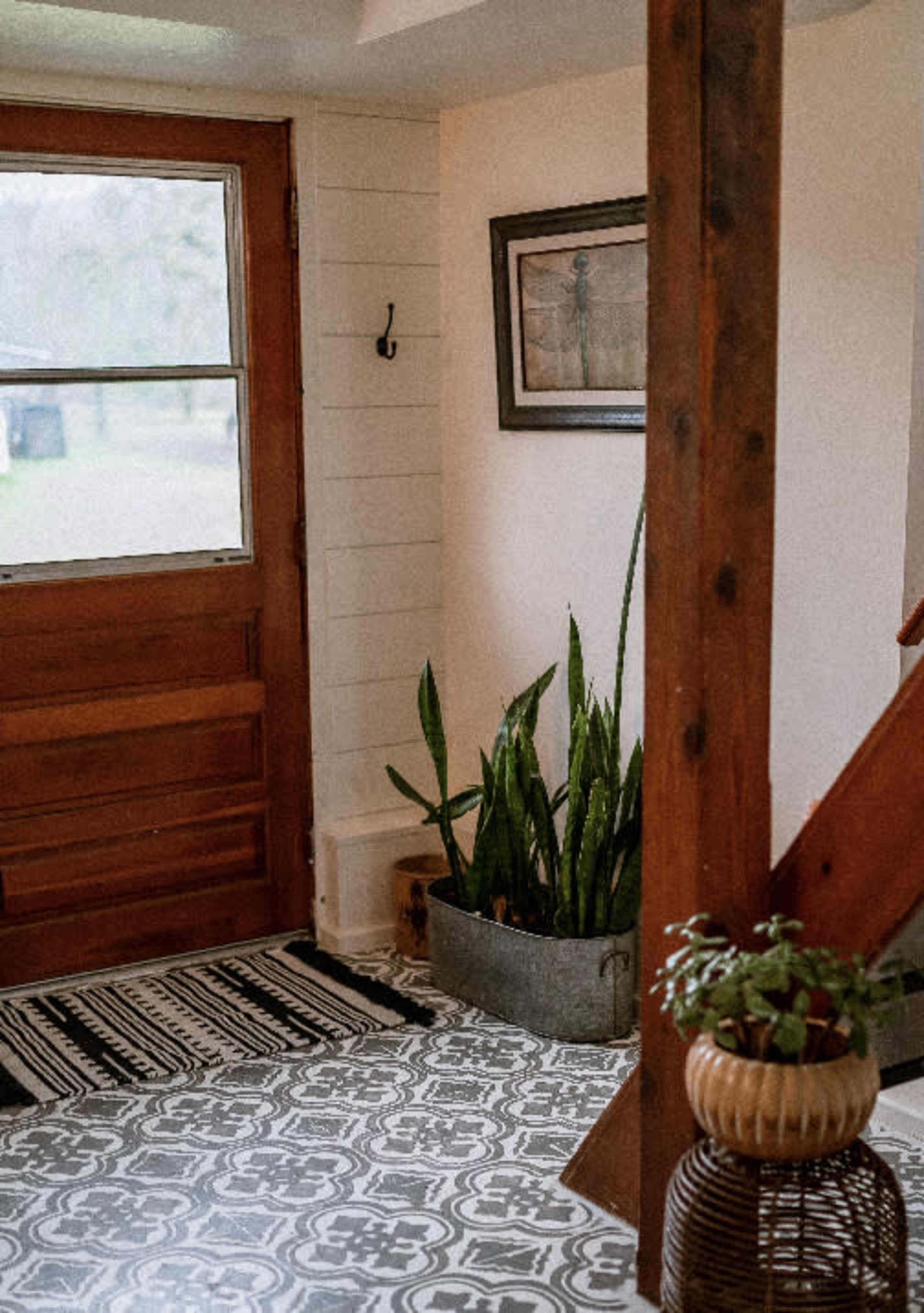The image shows an entryway with a wooden door, a patterned rug, a potted plant in a metal container, and a staircase.