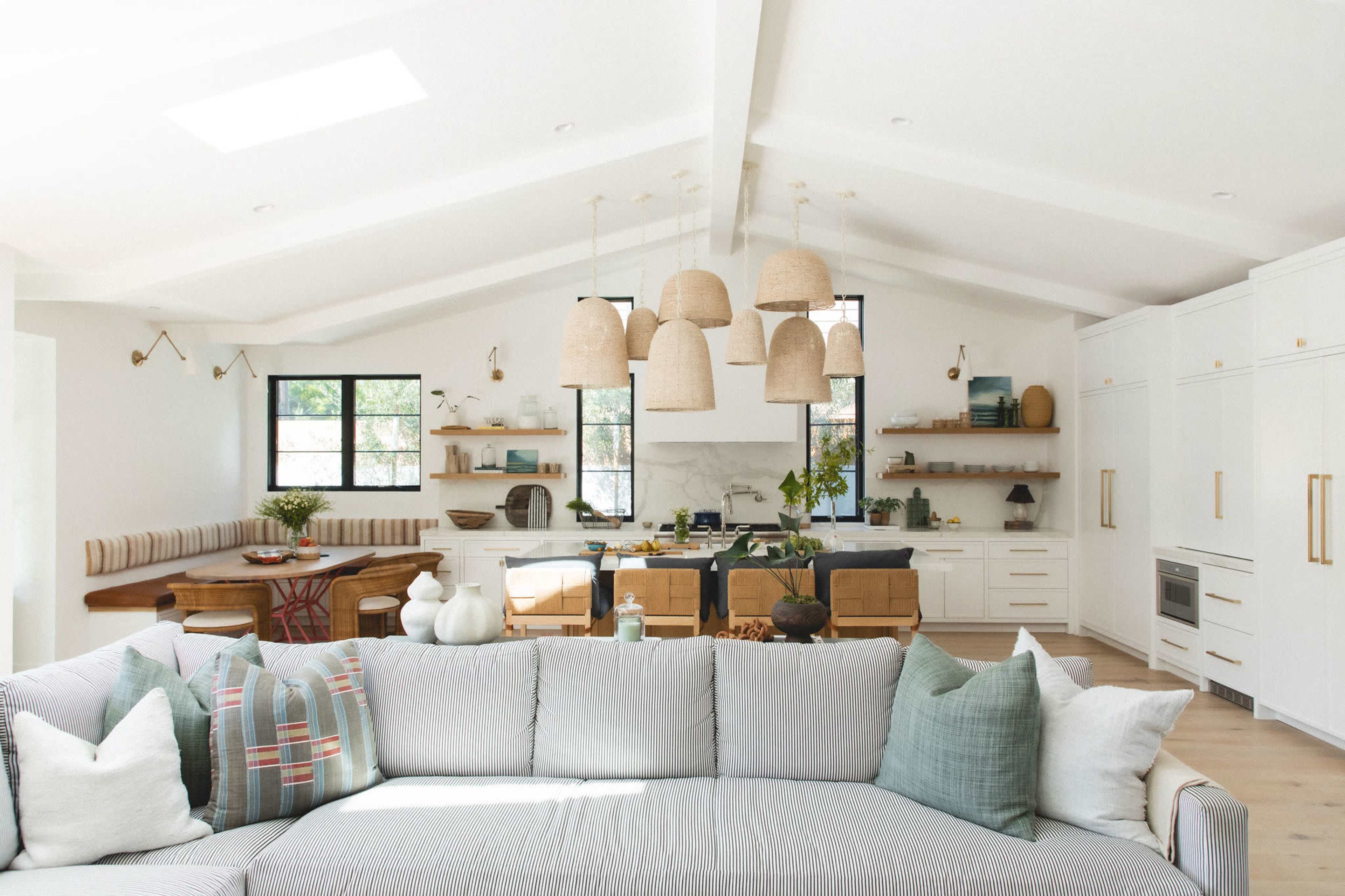 The image shows a modern, airy living space with a light-colored sofa in the foreground, a wooden dining table, and a kitchen area featuring minimalistic cabinetry and large pendant lights.