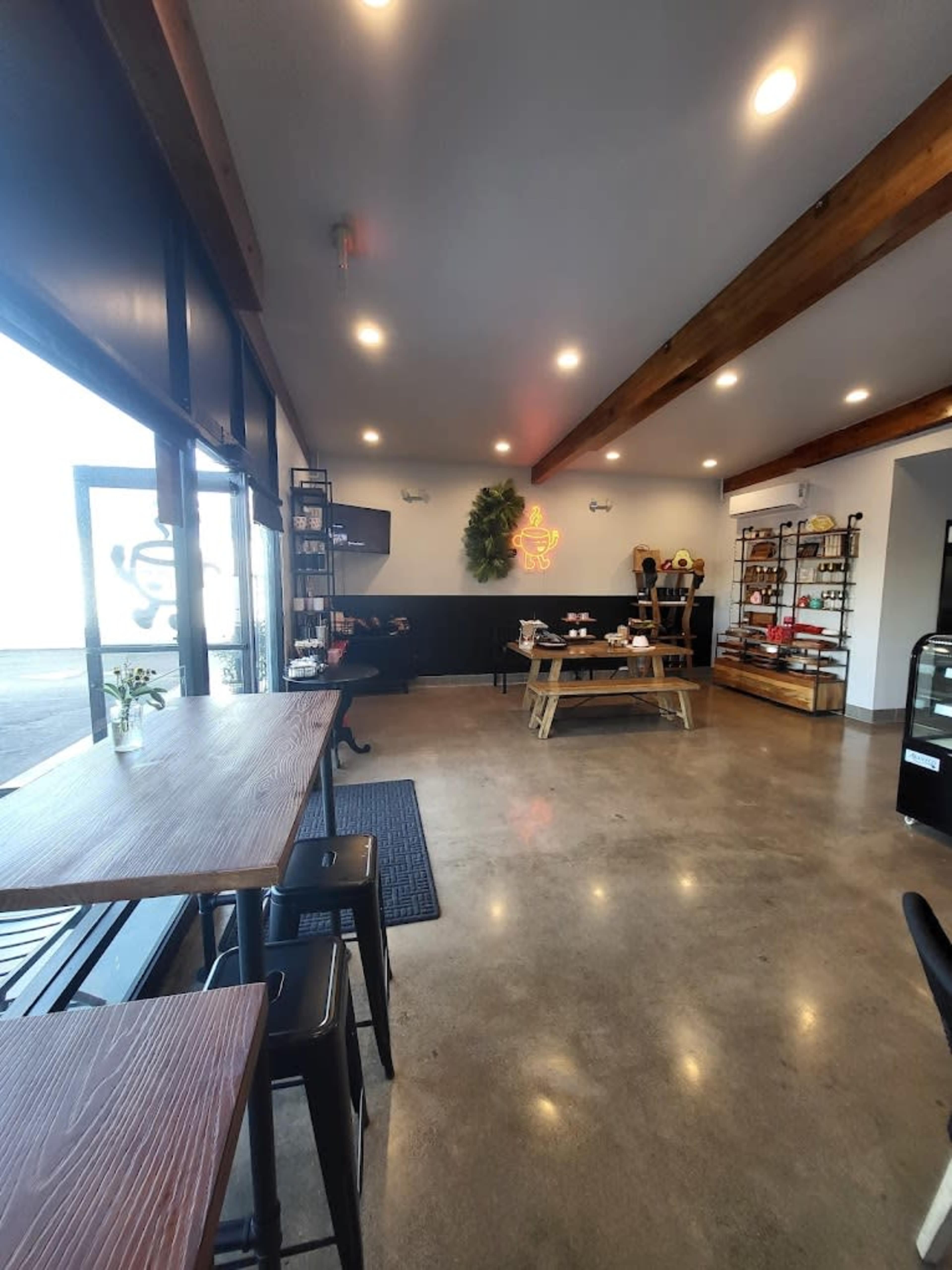 A modern cafe with wooden beams, concrete floors, and seating areas featuring tables and chairs, along with shelves displaying various products.