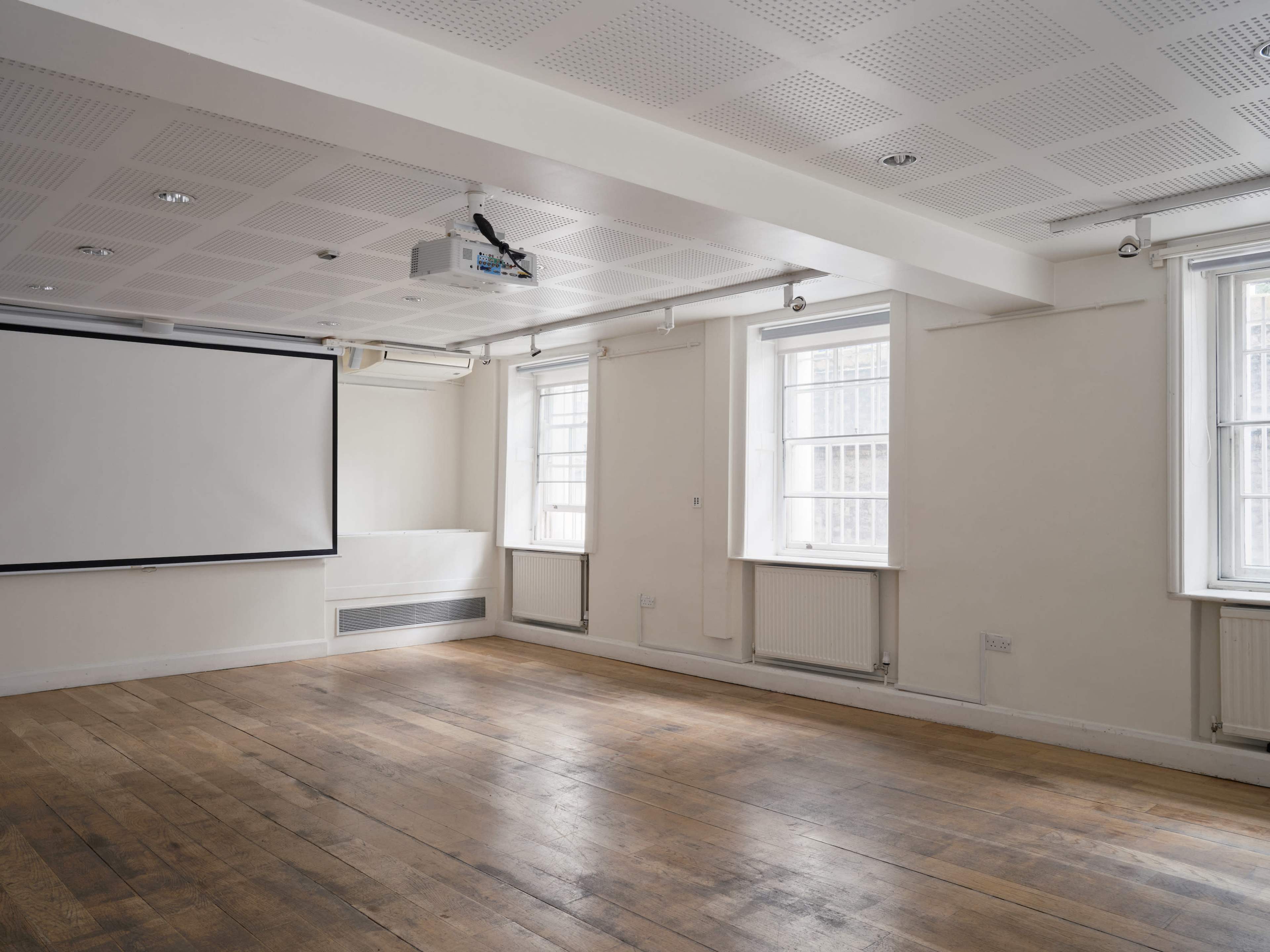 The image shows a spacious, empty room with wooden flooring, a projector hanging from the ceiling, and a large screen mounted on the wall.