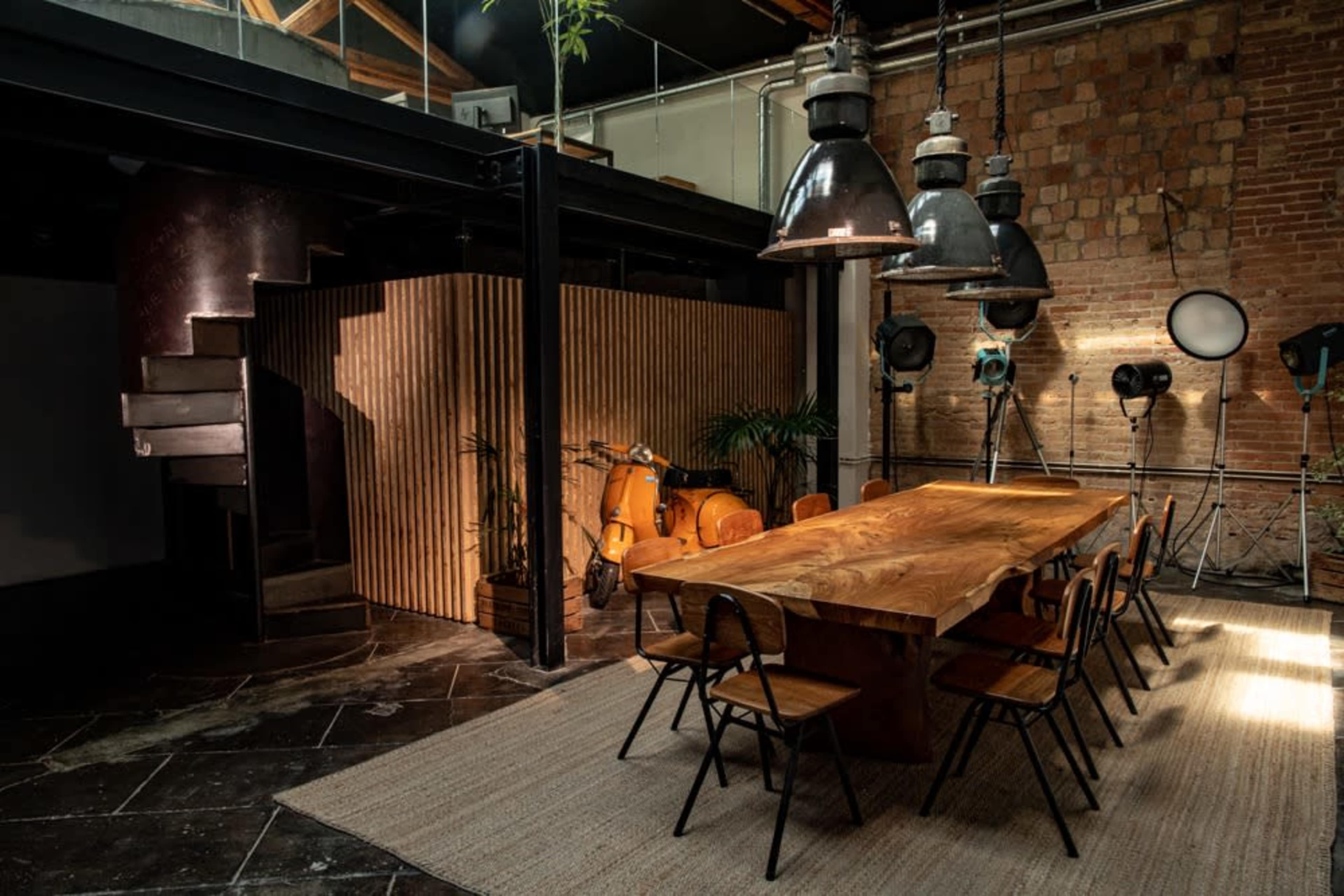 A spacious, modern interior featuring a large wooden dining table surrounded by black chairs, with industrial lighting and a staircase in the background.