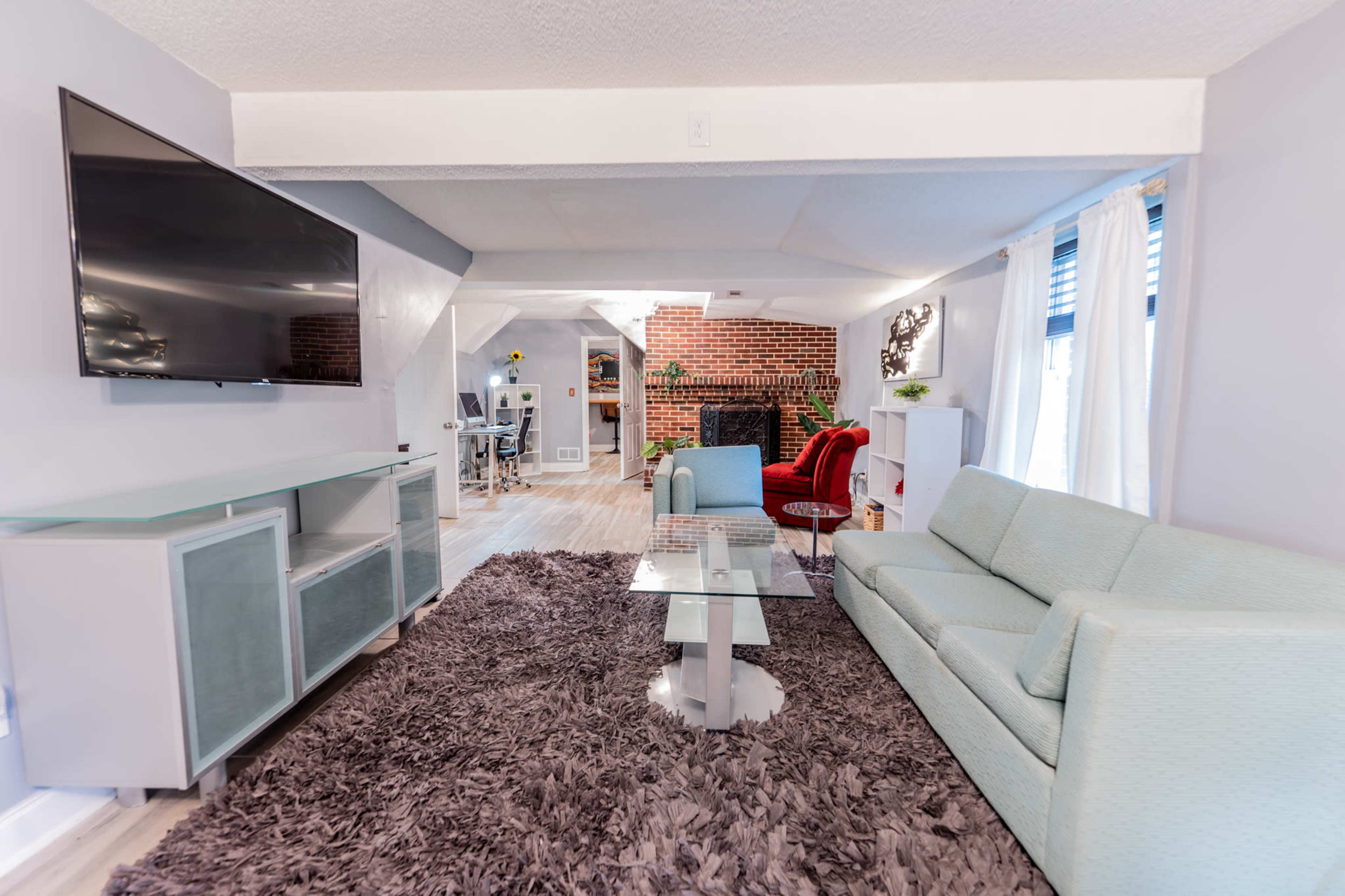 The image shows a modern living room with a light blue sofa, a glass coffee table, a large flat-screen TV, and a textured rug, featuring a brick fireplace in the background.