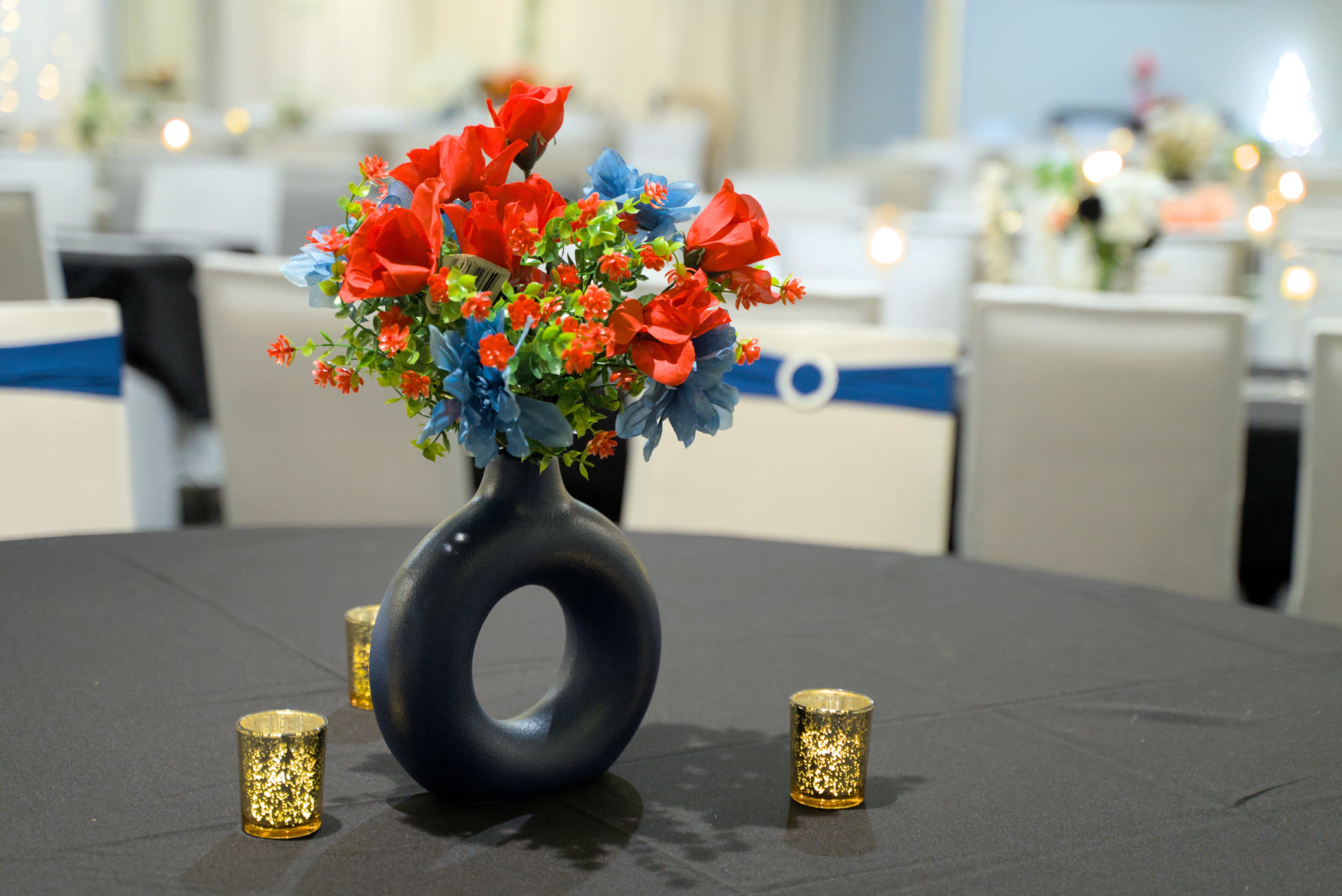 A black table covered with a cloth features a circular vase filled with red and blue flowers, surrounded by glittering candle holders.