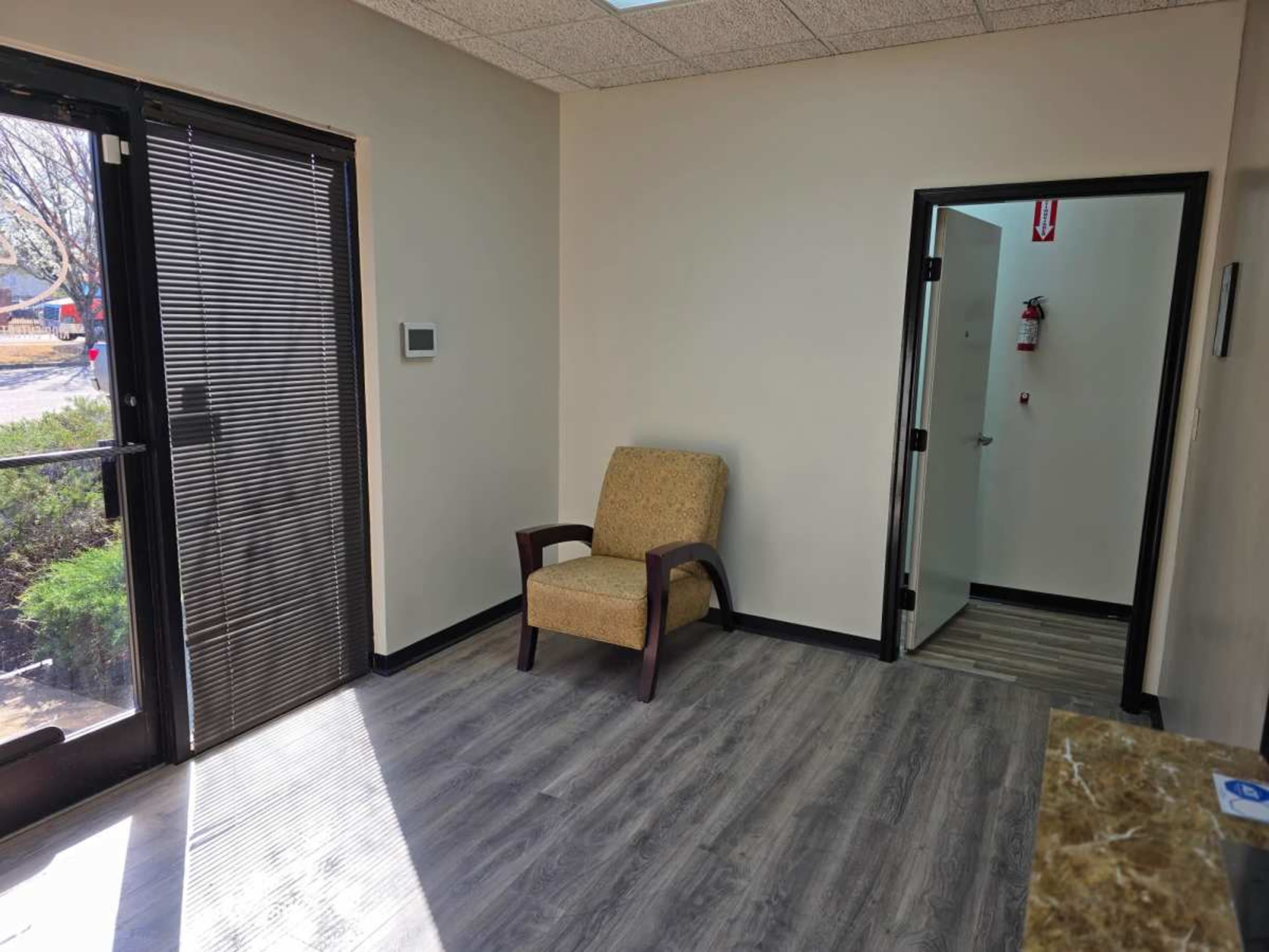 The image shows a small waiting area with a single chair, a door leading to another room, and large glass doors that open to the outside.
