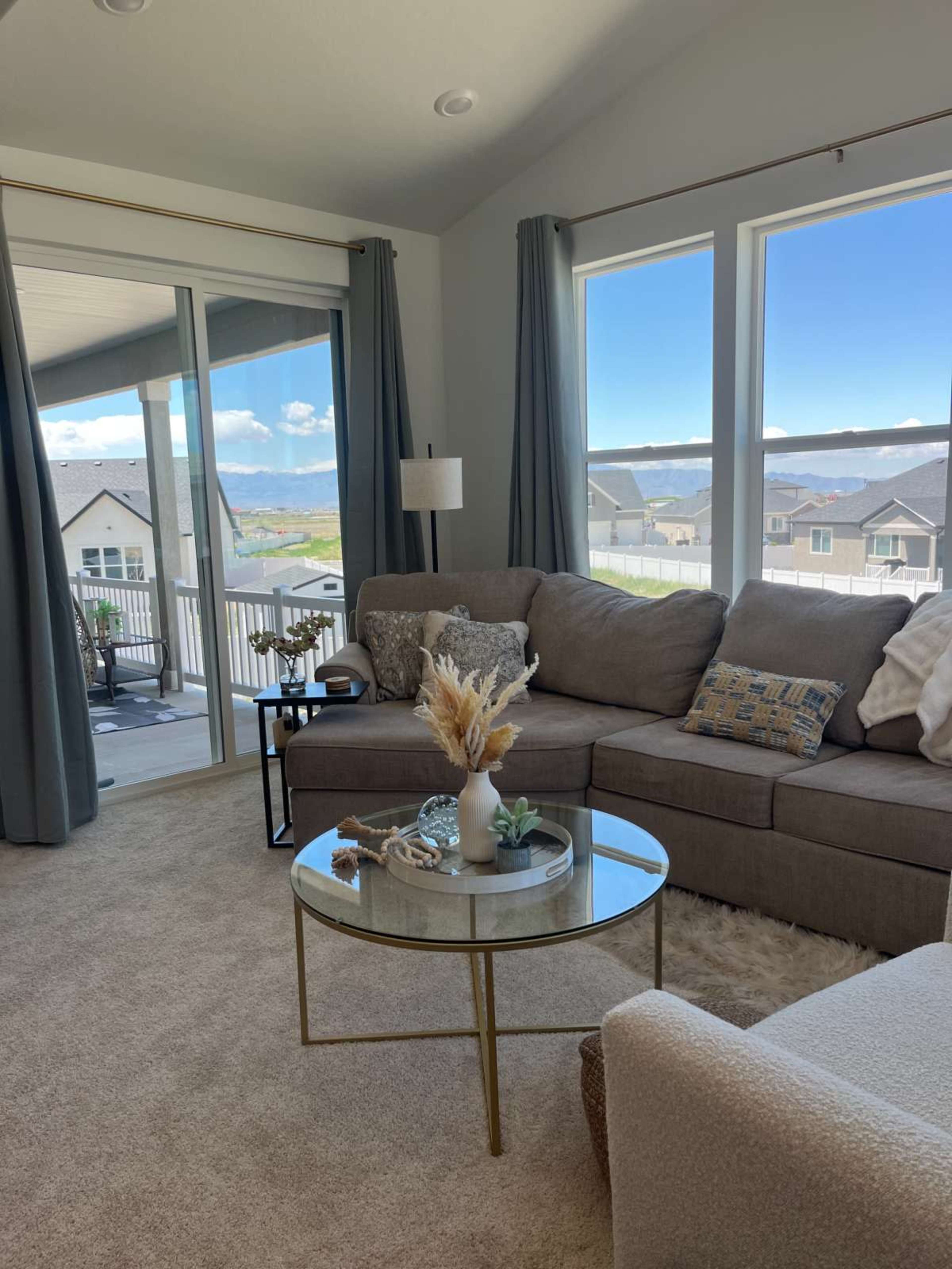 The image shows a cozy living room with a sectional sofa, a small round coffee table, and large windows offering a view of the outdoors against a backdrop of mountains and houses.