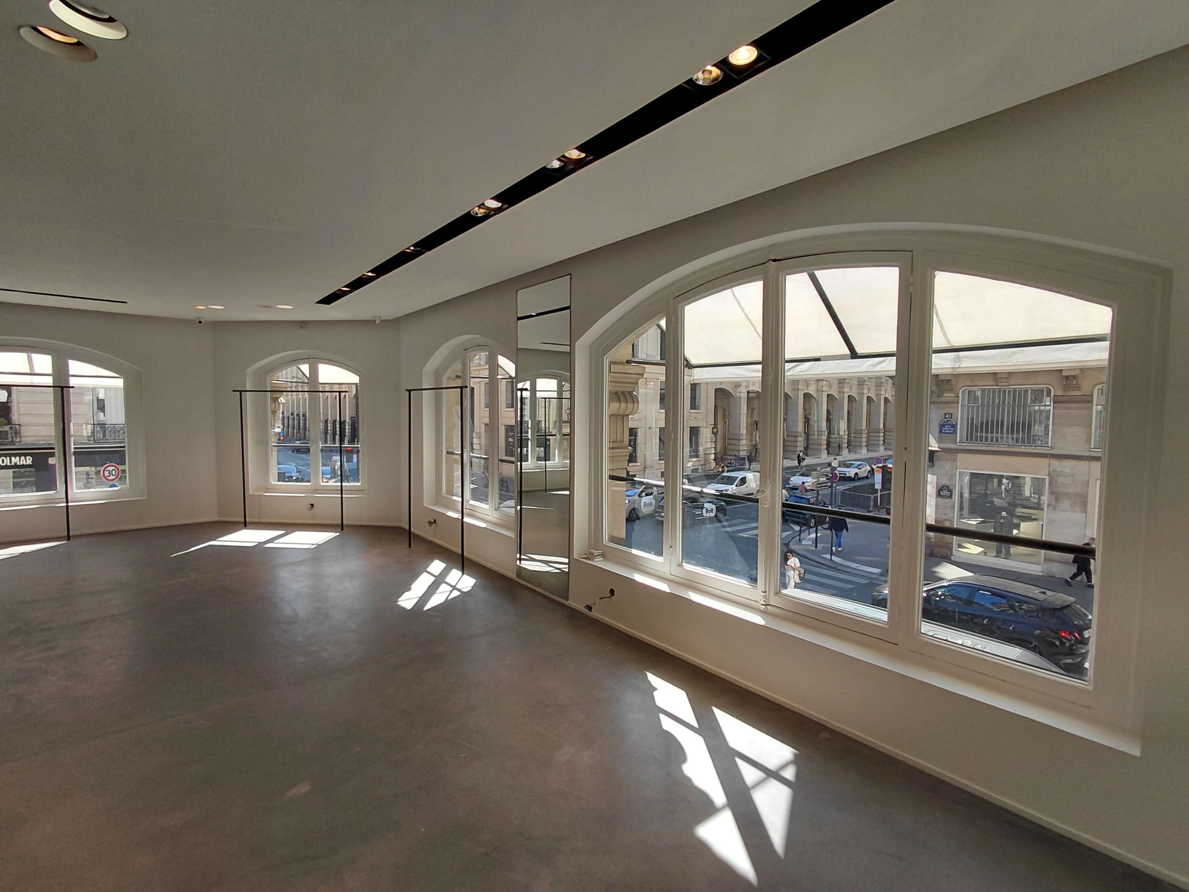 The image shows a spacious, empty interior with large arched windows allowing natural light to enter, revealing a view of a street lined with cars and people outside.