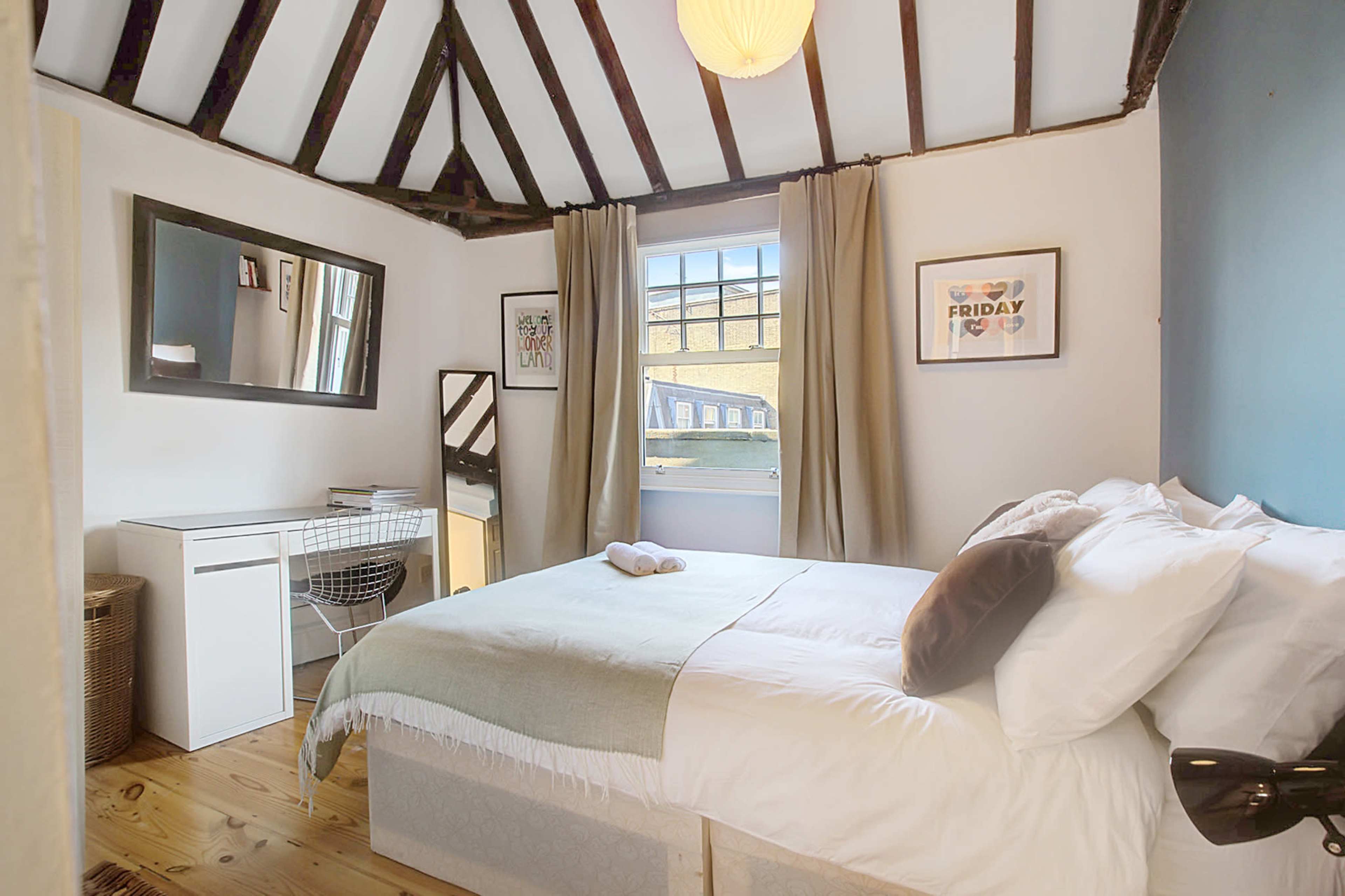 The room features a bed with white bedding, a small desk and mirror, and natural light from a window with curtains.