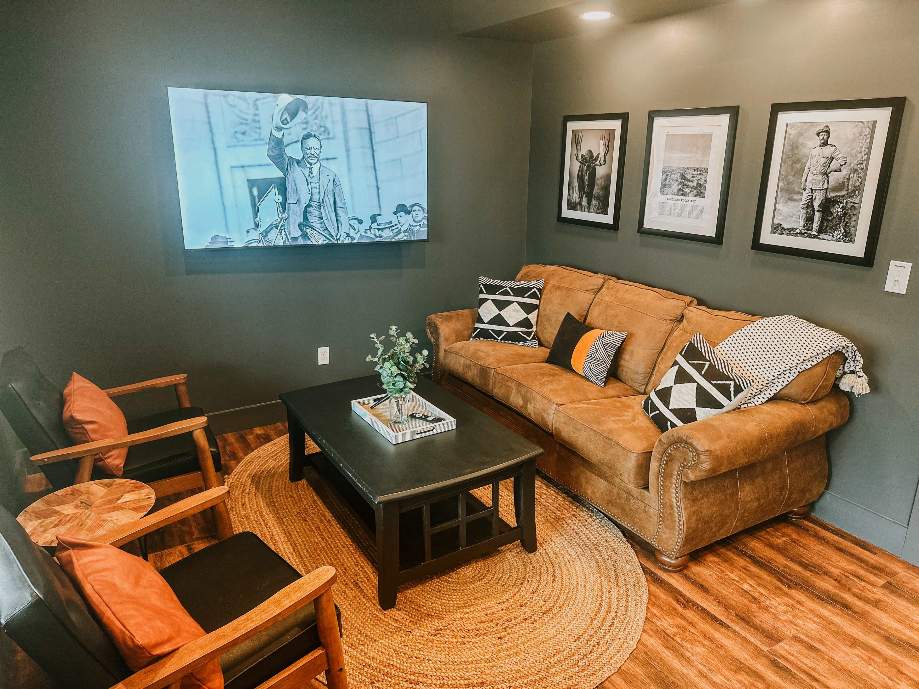 A cozy living room with a brown sofa, two wooden chairs, a black coffee table, and framed photos on the walls, complemented by a large television displaying black and white images.