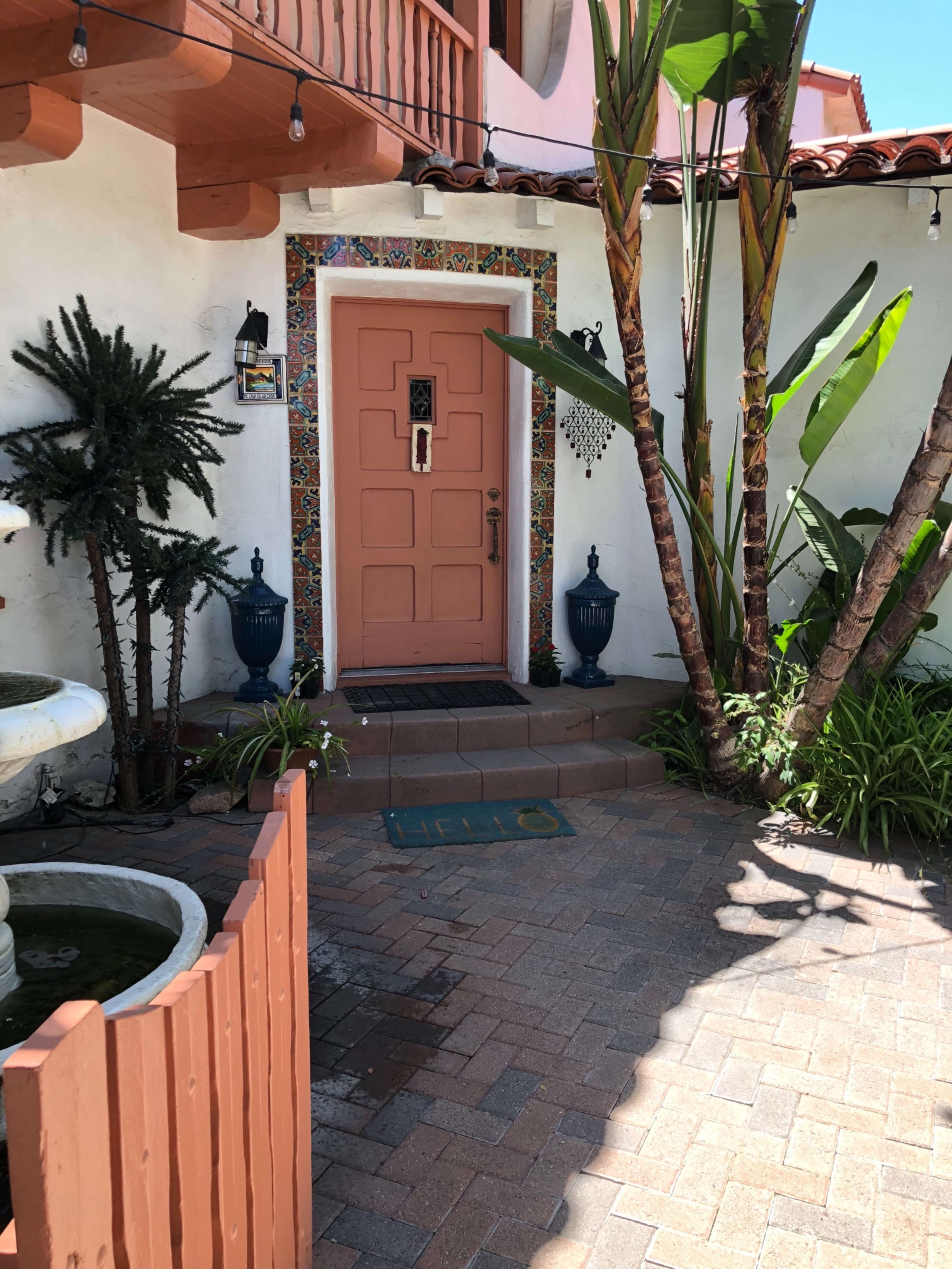 The image shows a pink front door surrounded by colorful tiles, flanked by tall green plants, with a pathway leading to it.