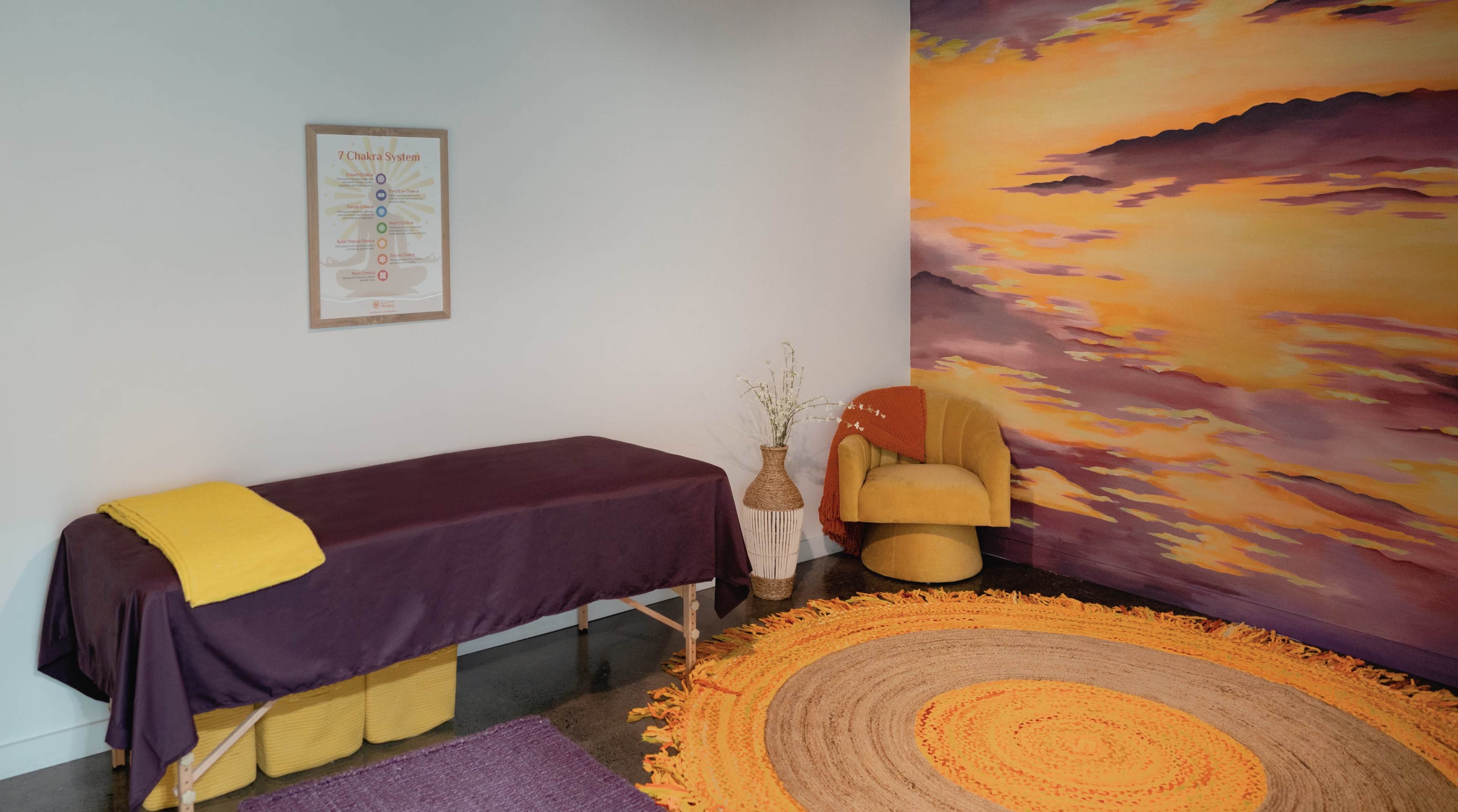 The image shows a wellness room featuring a massage table draped in a purple cover, a colorful mural of clouds on the wall, and a yellow armchair beside a woven rug.