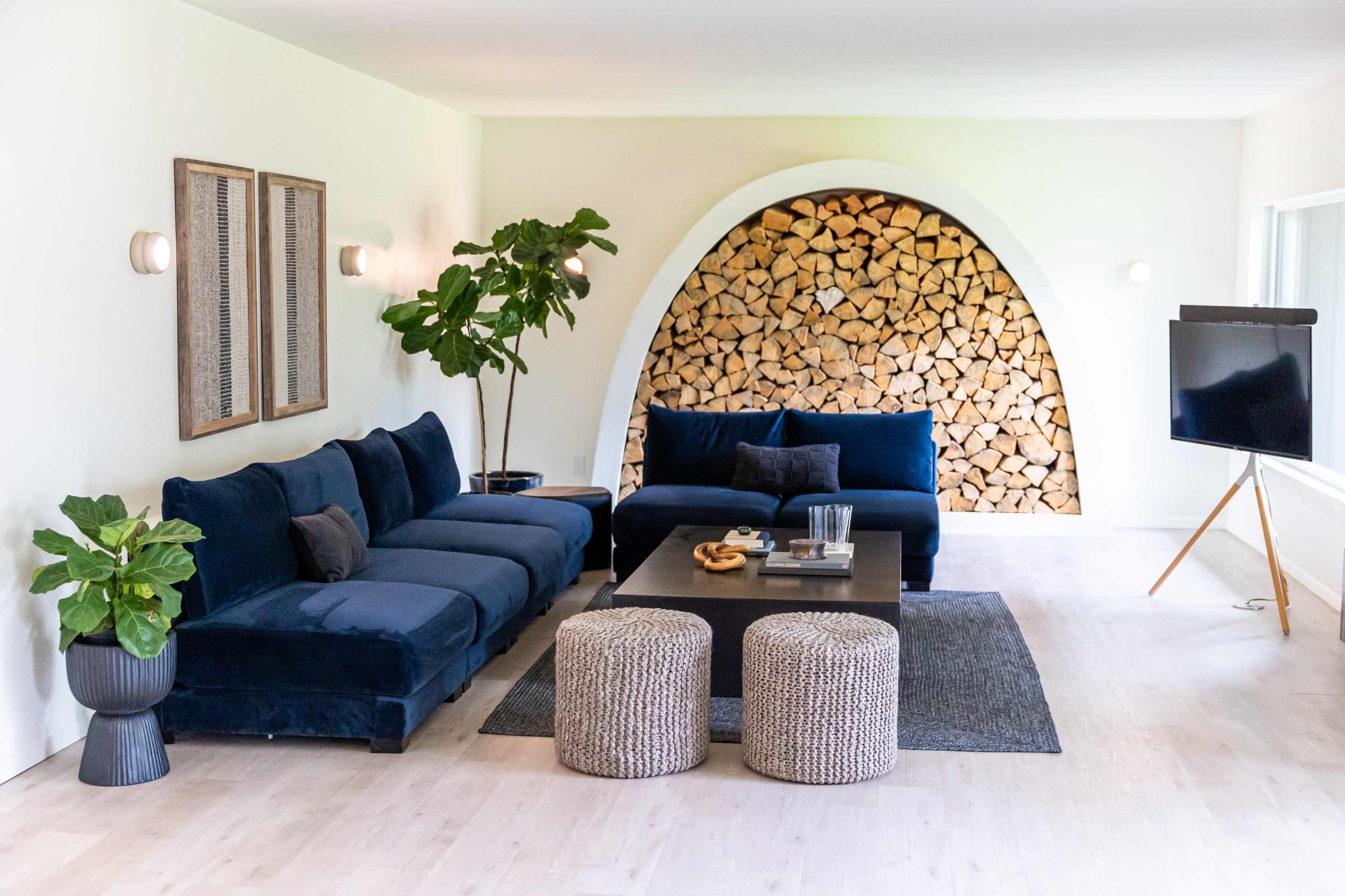 A modern living room featuring a blue sectional sofa, a round stack of firewood in an archway, and a coffee table with two textured poufs.