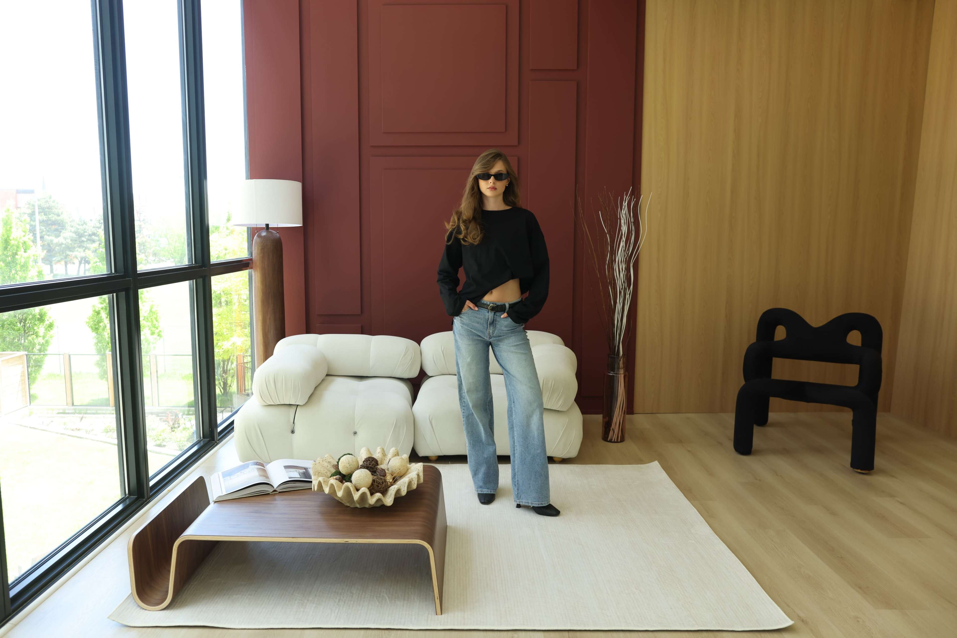 A woman with long hair wearing sunglasses stands in a modern living room with large windows, a sculptural coffee table, and minimalist furniture.