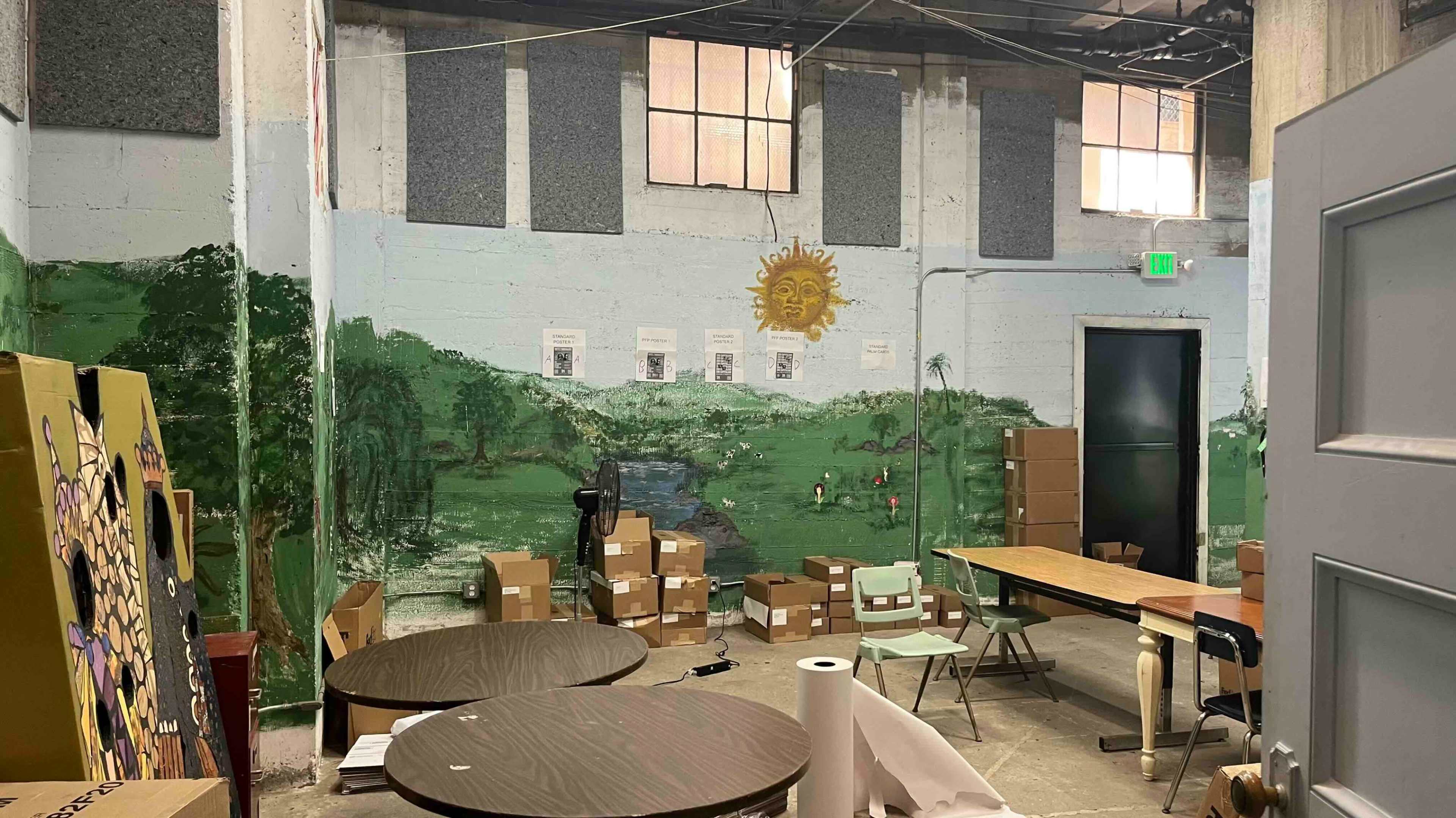 The image shows a partially furnished room with a mural of a landscape on the wall, surrounded by cardboard boxes and tables.
