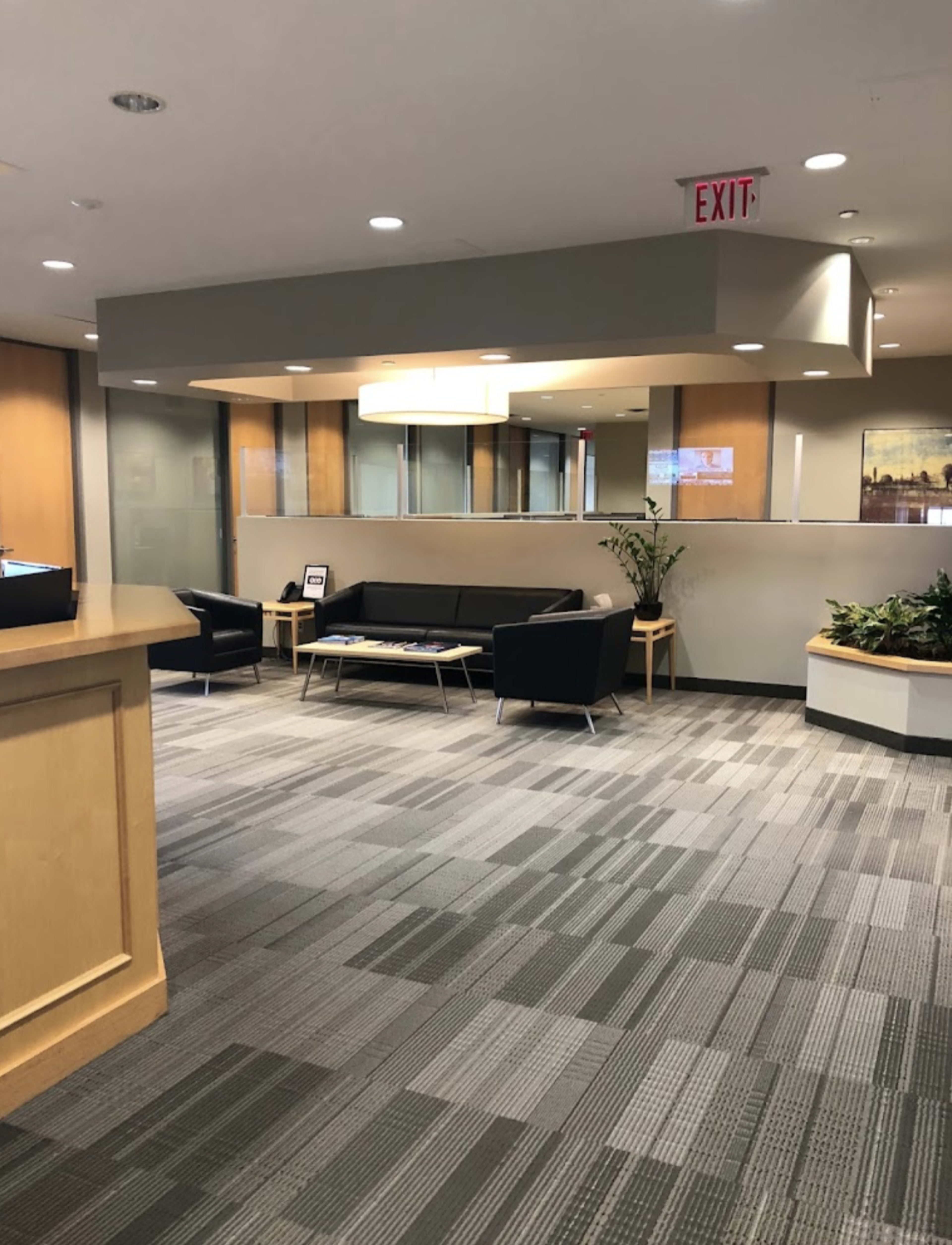 An office reception area features a seating arrangement with a black couch and chairs, along with plants and a large window in the background.