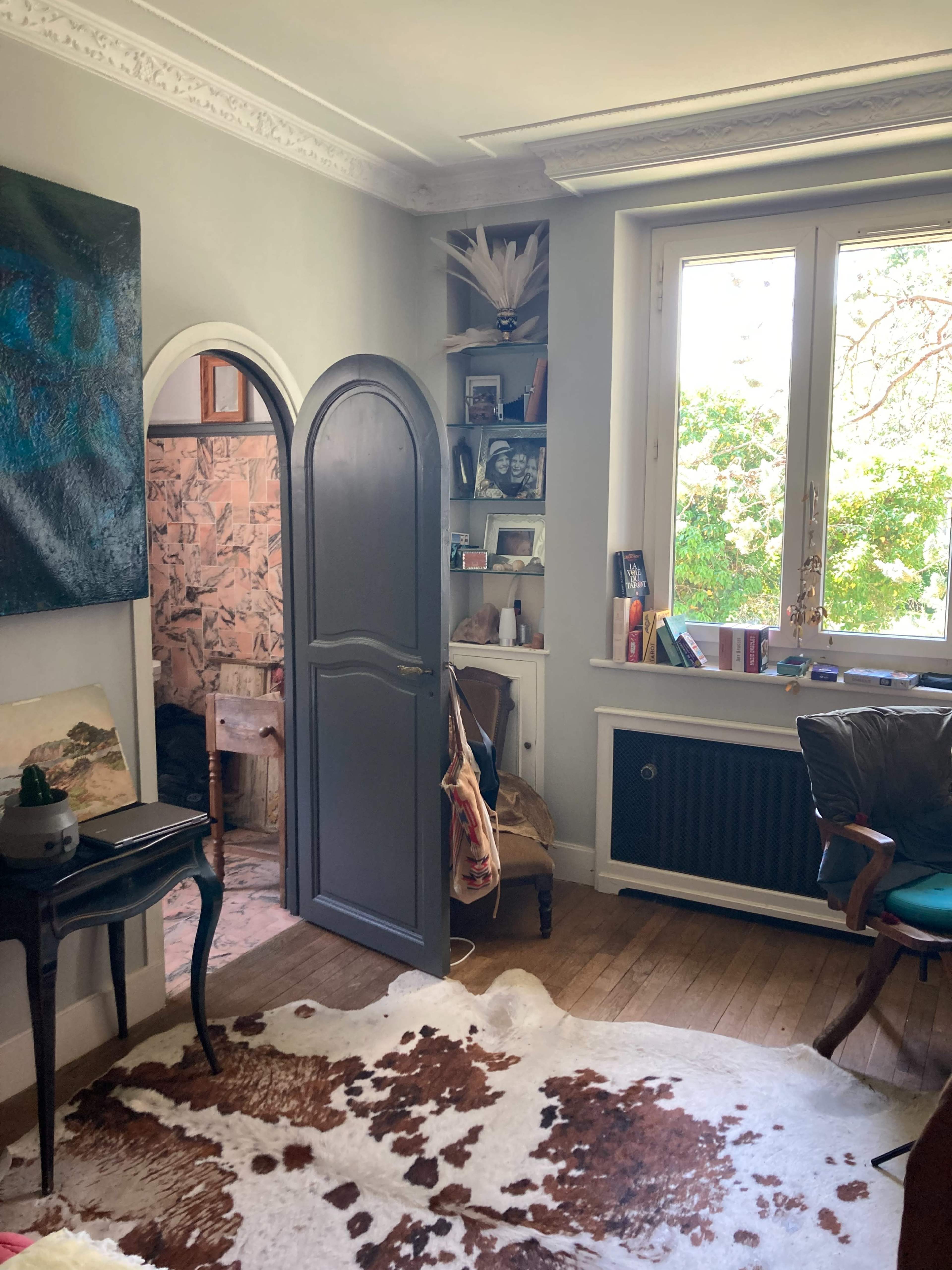 A cozy room features a dark arched door leading to another space, a bookshelf filled with framed photos, and a large window offering a view of greenery outside.