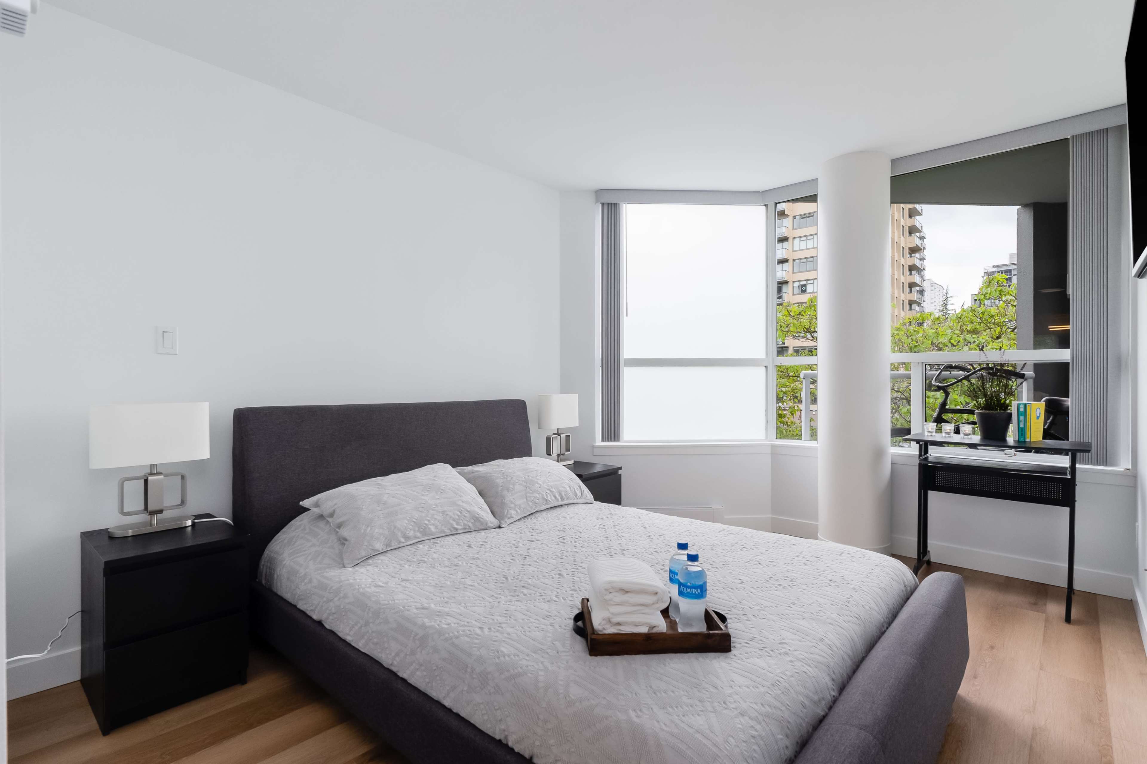 A modern bedroom features a gray bed with white linens, two lamps on either side, a small desk, and large windows overlooking greenery.