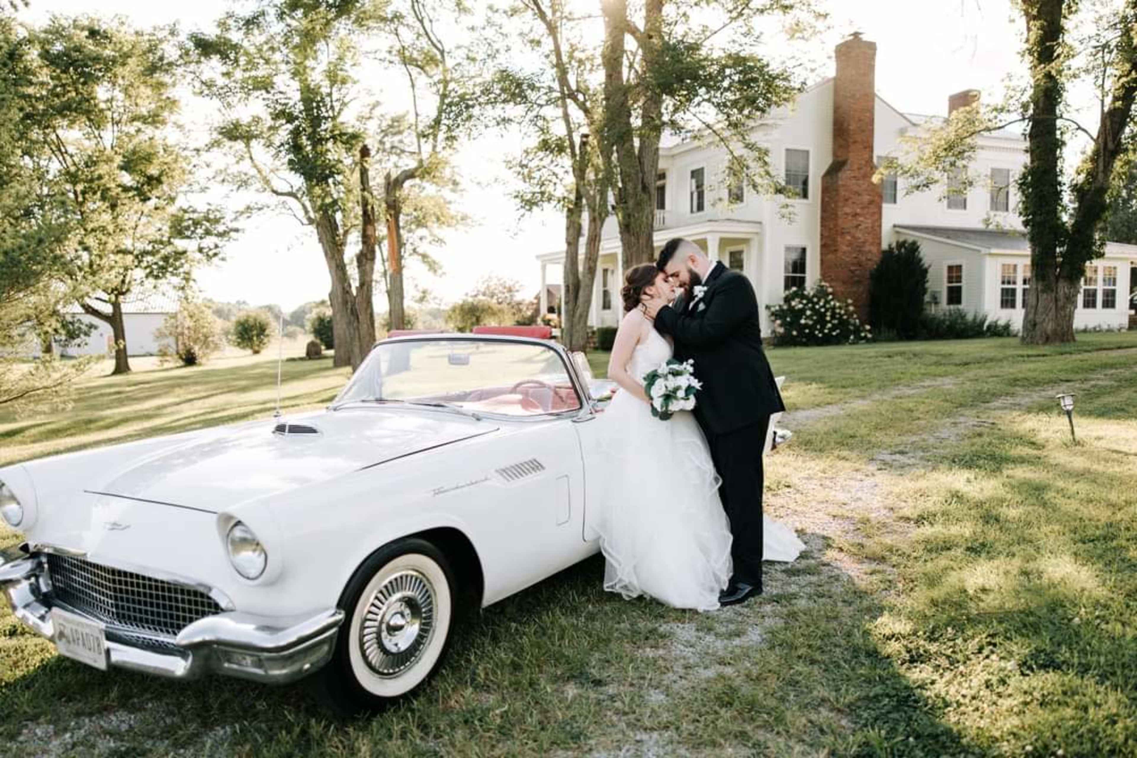 A couple in formal attire embraces near a white vintage car in front of a large house surrounded by trees.