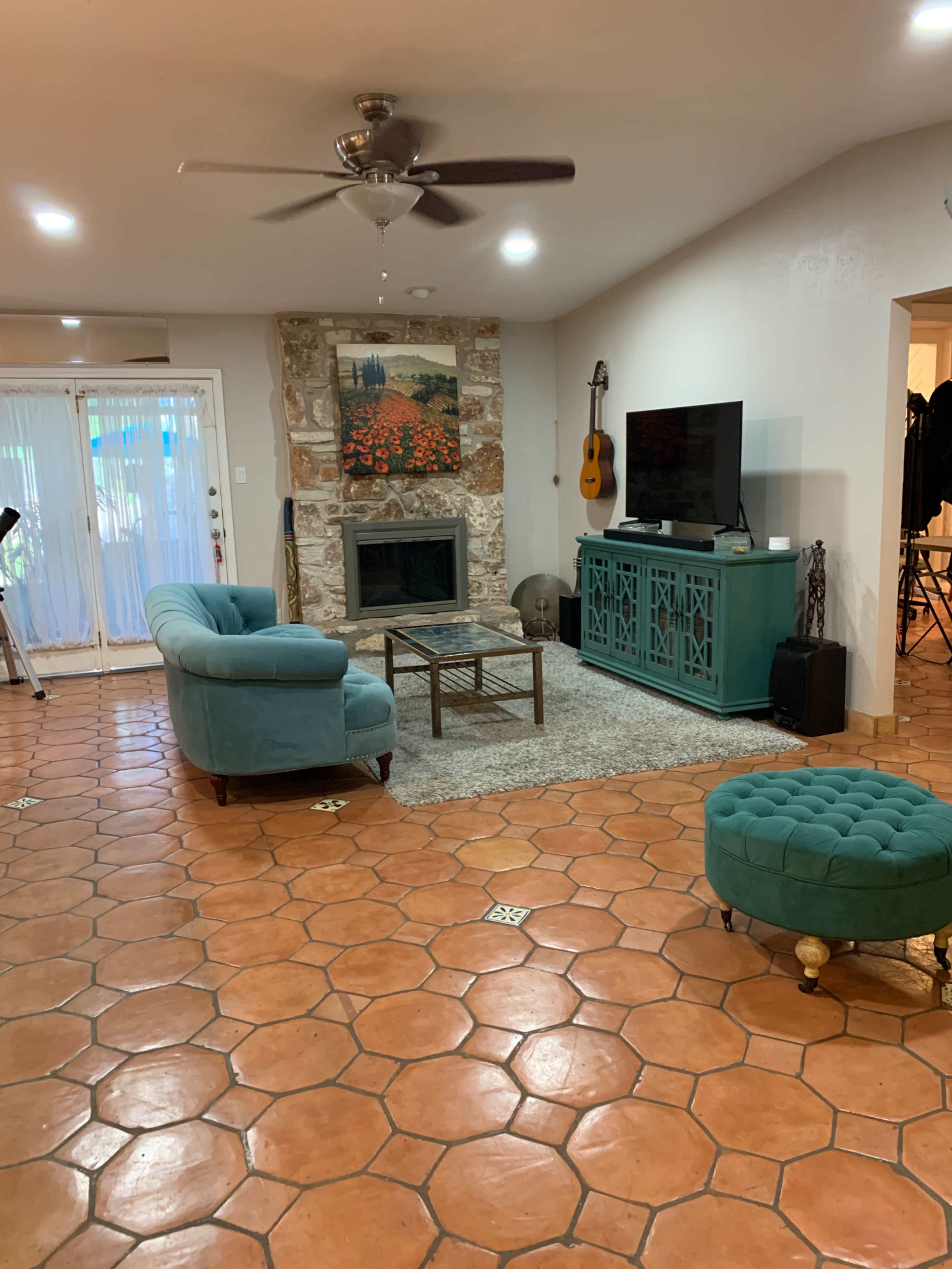 The living room features a turquoise couch and matching ottoman, a stone fireplace, and a television mounted on a decorative cabinet, all set against terracotta tile flooring.