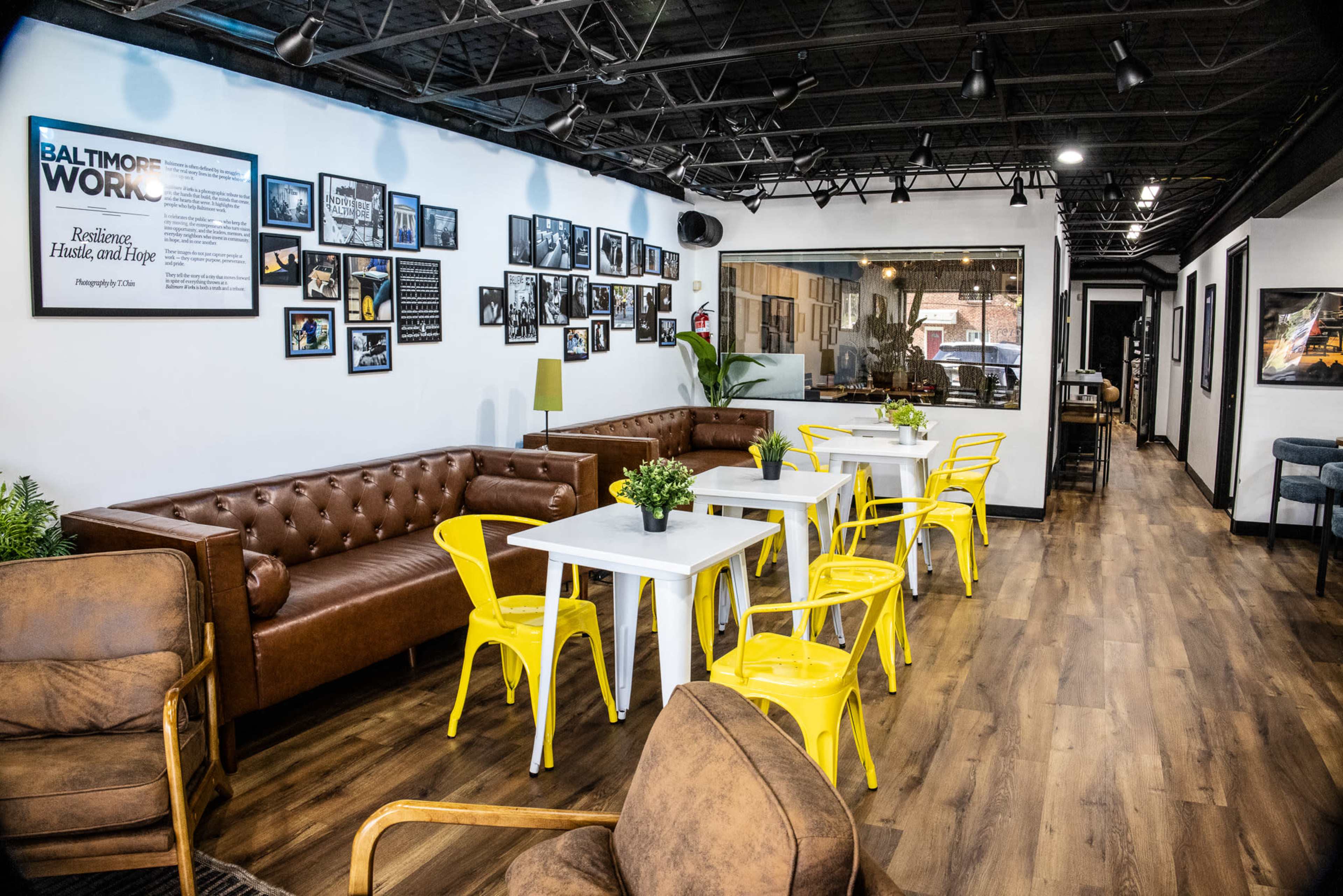 The image shows a modern café interior with brown leather sofas, yellow chairs around white tables, and framed photographs on the walls.