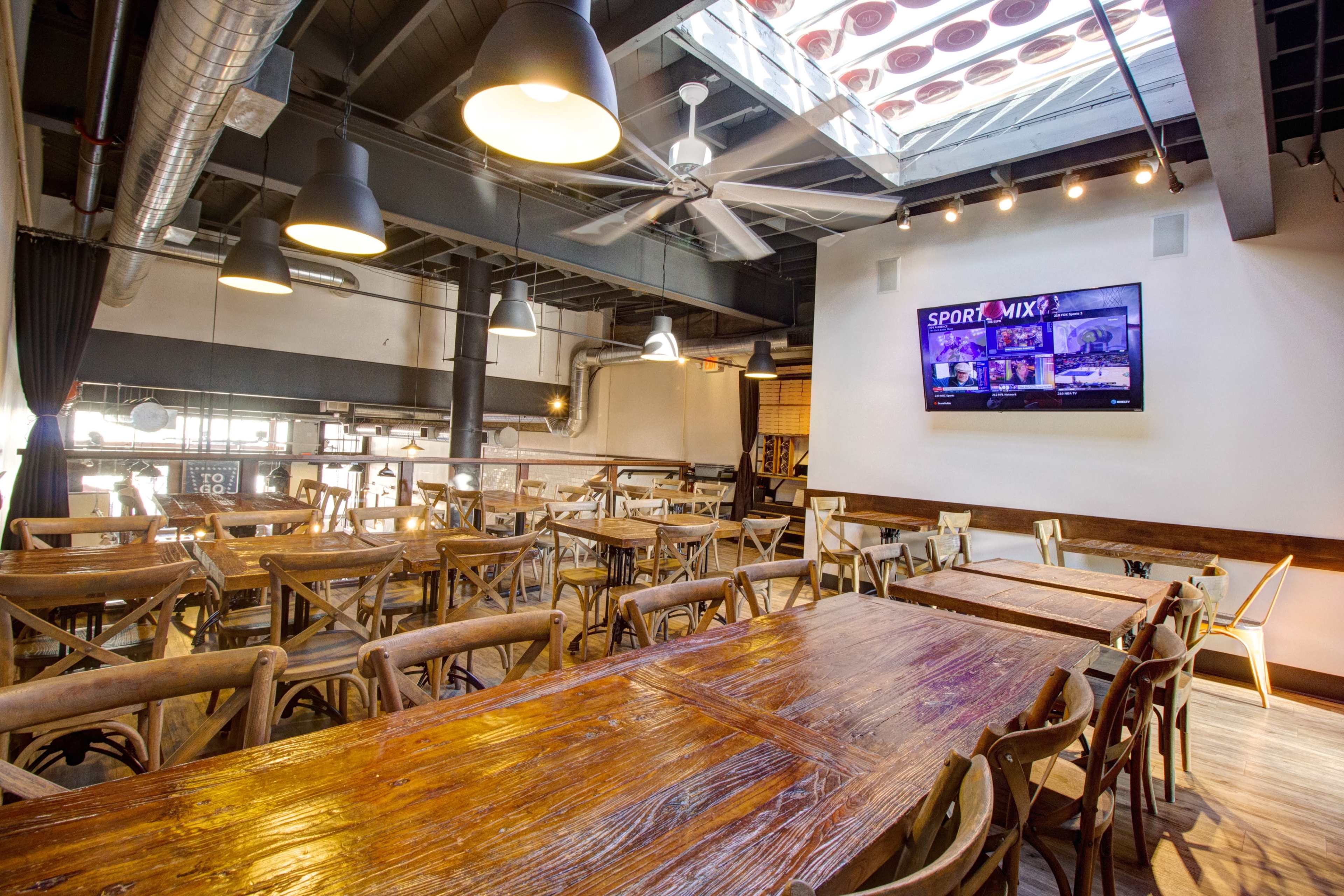 The image shows a spacious, modern restaurant interior with wooden tables, overhead fans, and a large screen displaying a sports channel.