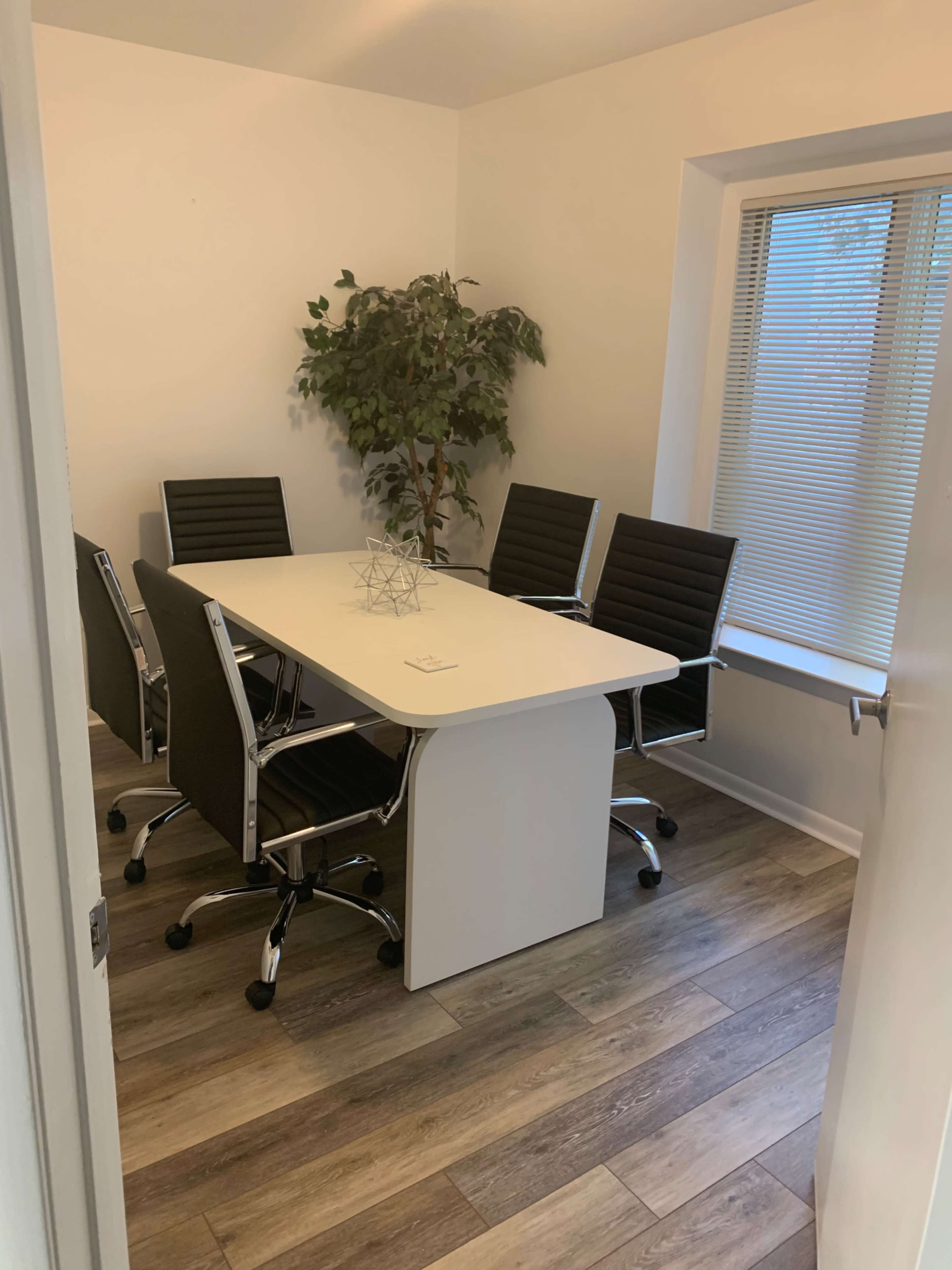 A modern conference room features a large white table surrounded by five black rolling chairs and a potted plant in the corner.