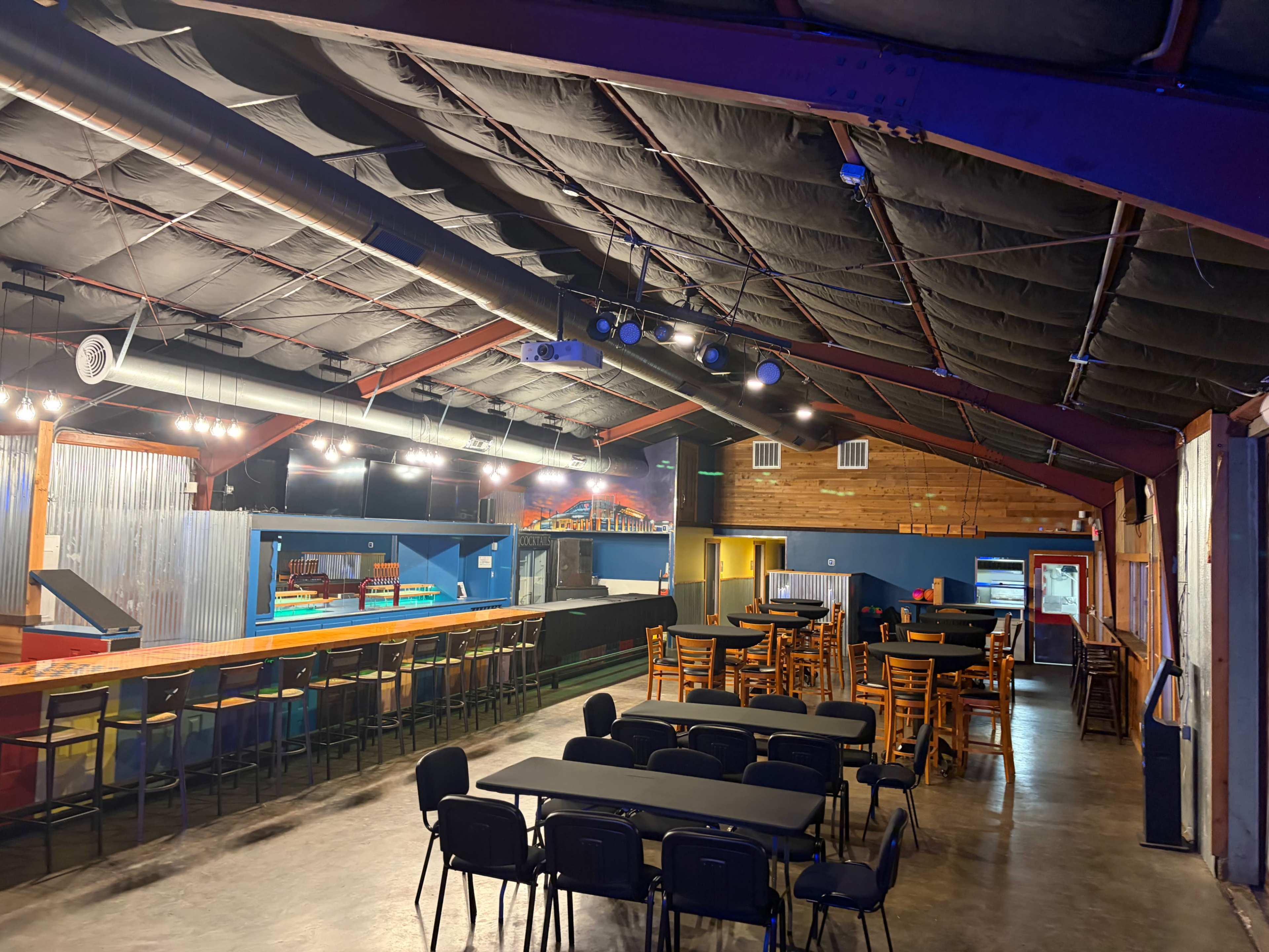 The image shows the interior of a large venue with a bar counter, multiple tables and chairs, and a stage area in the background.