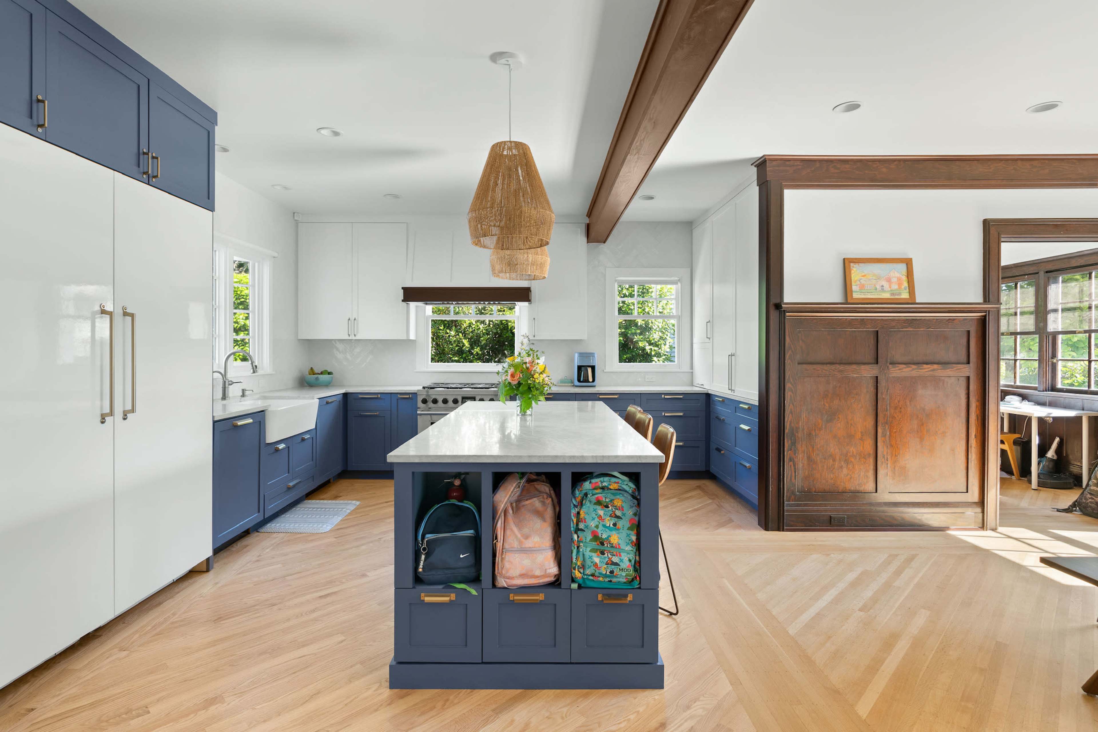 A modern kitchen features a central island with blue cabinetry and a marble countertop, complemented by white upper cabinets and a wood beam ceiling.