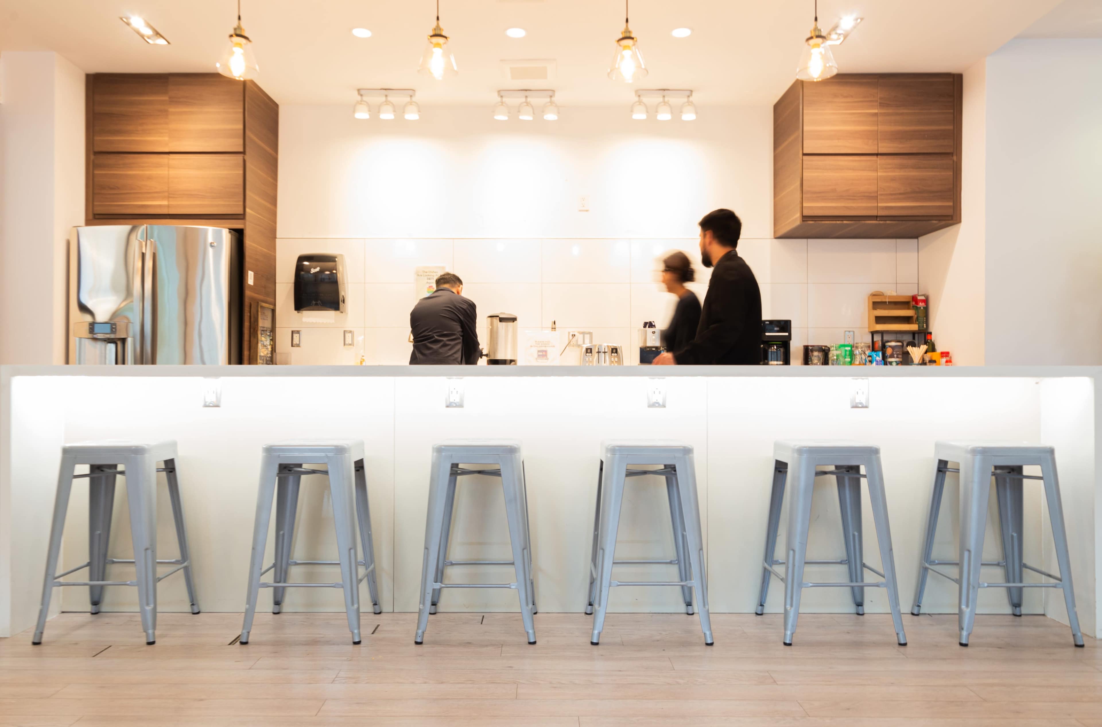A modern kitchen with a white countertop and metal stools, featuring a person preparing coffee in the background under pendant lighting.