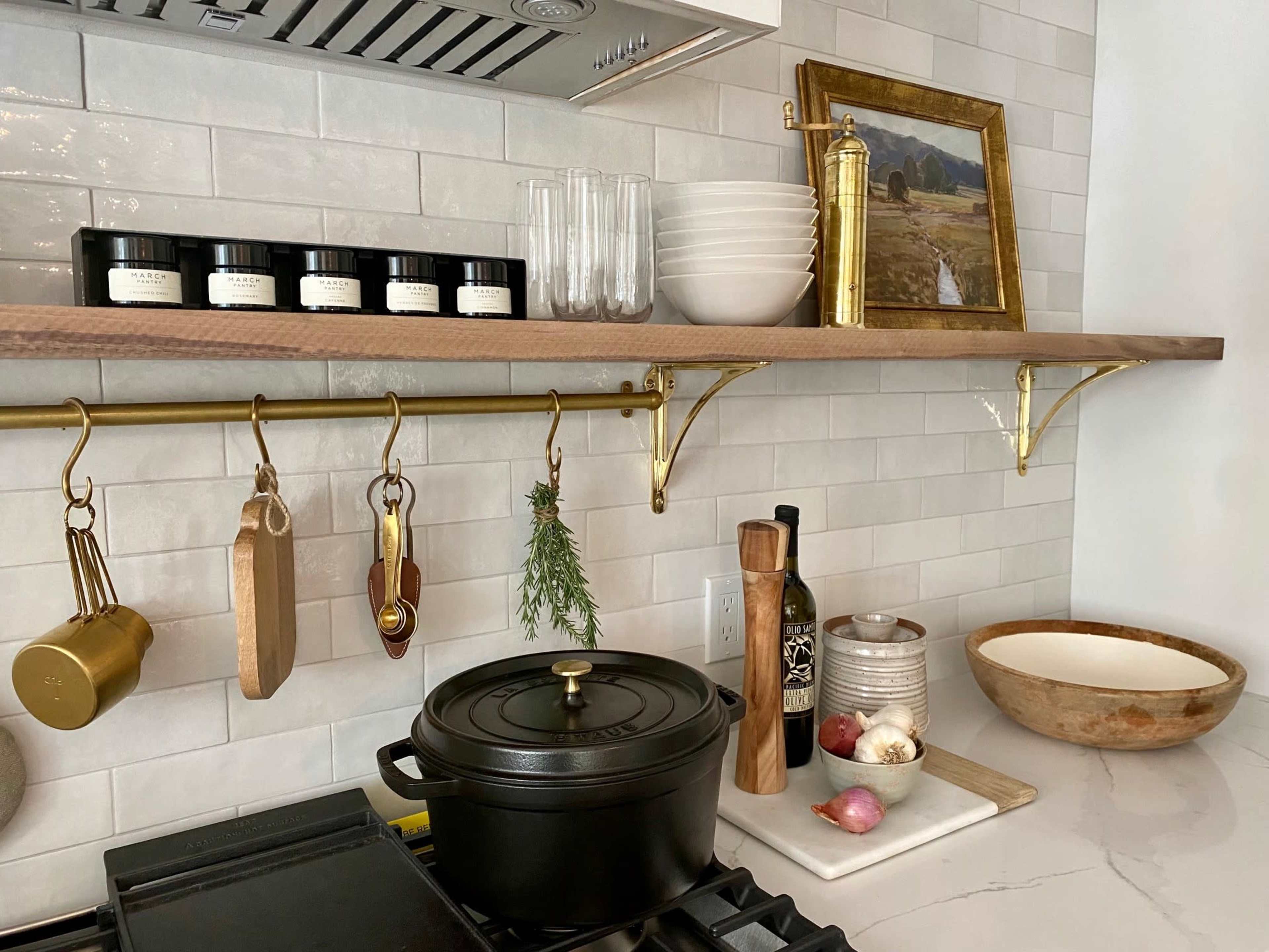 A kitchen countertop displays a cast-iron pot, various cooking utensils, and decorative items on a wall-mounted shelf with brass brackets.