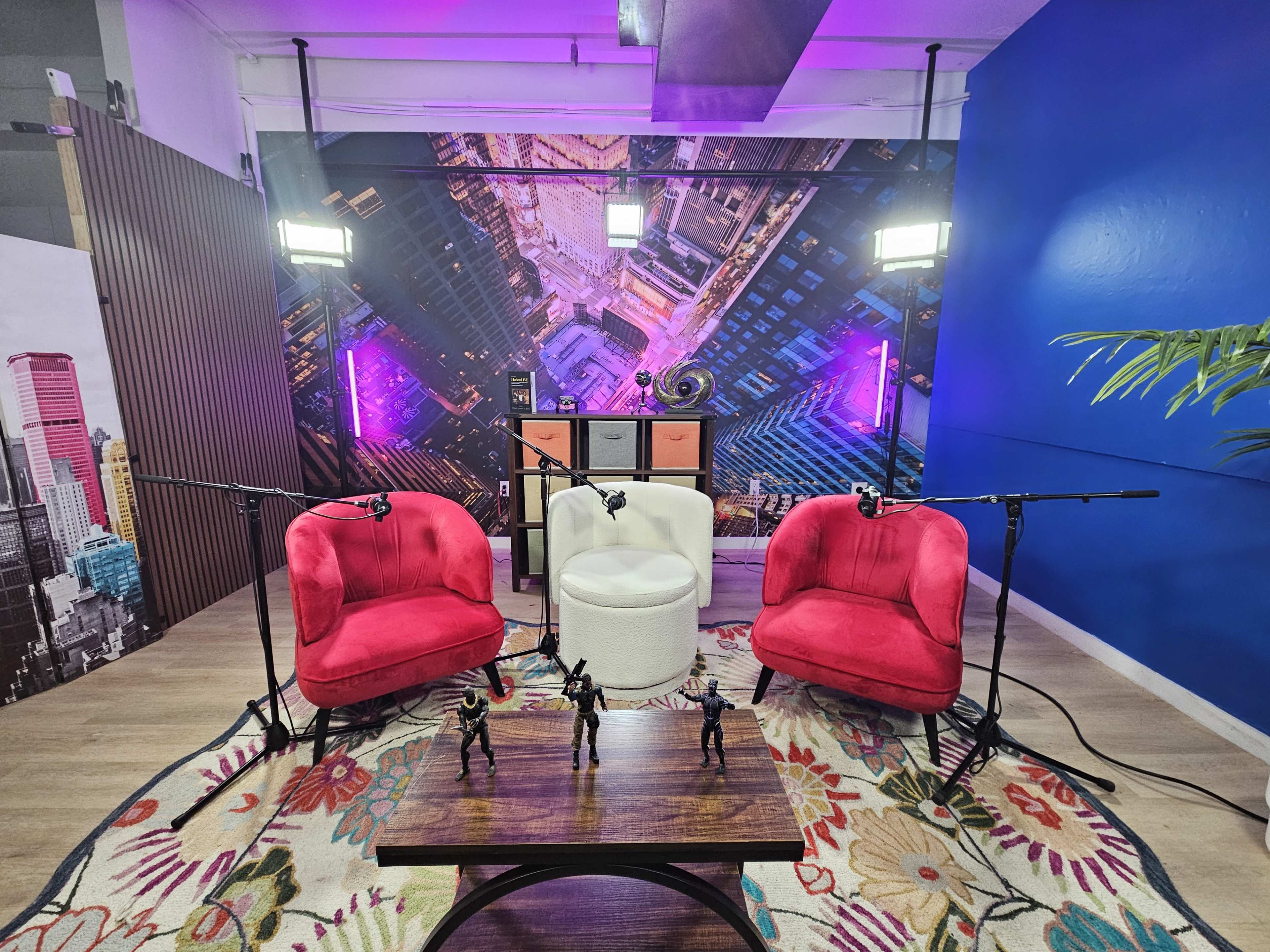 The image shows a modern studio setup with two pink chairs, a white stool, and a dark wooden table, surrounded by colorful lighting and a cityscape backdrop.