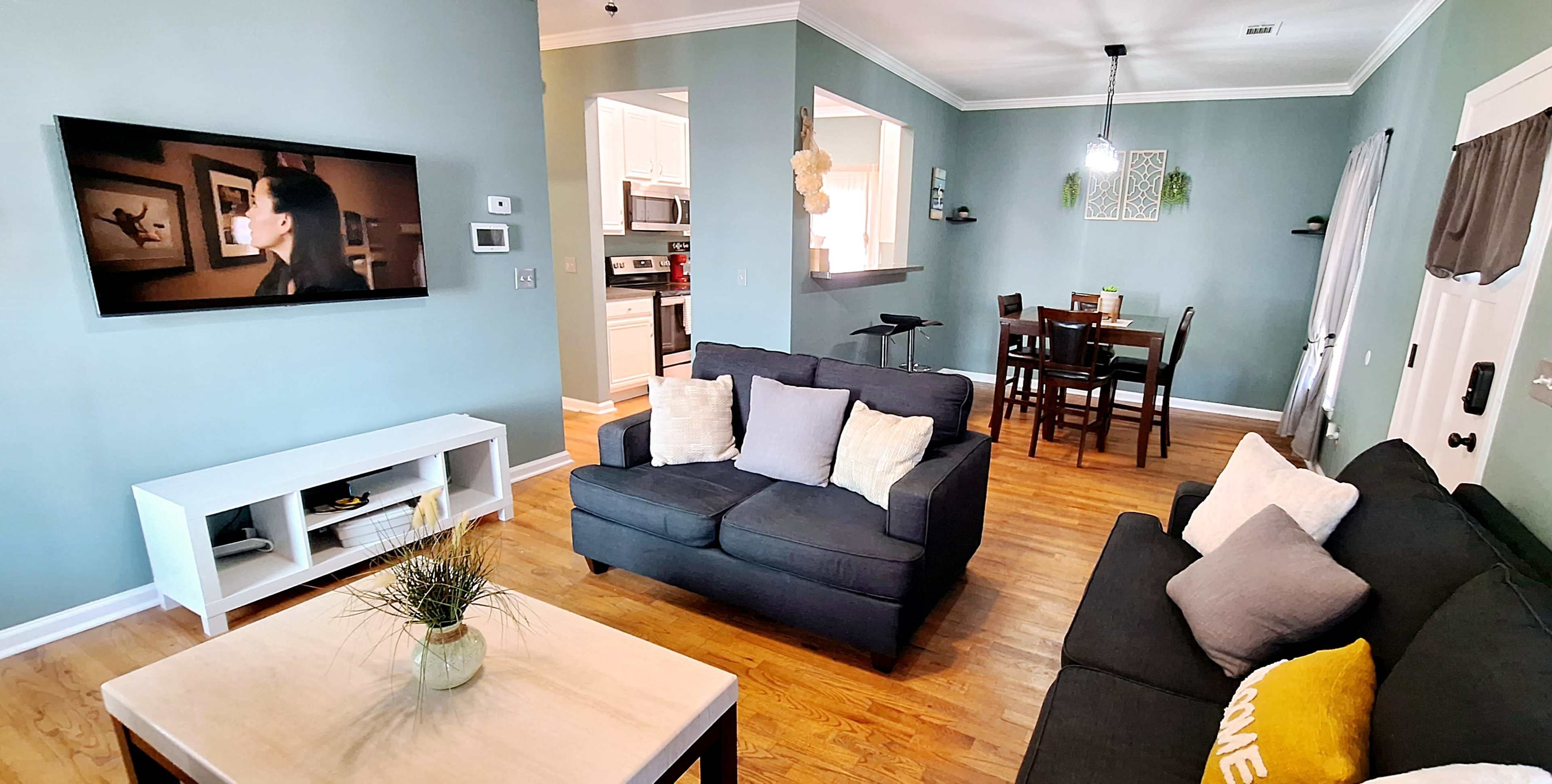 A living room with a dark sofa facing a television, adjacent to a dining area and a kitchen visible in the background.